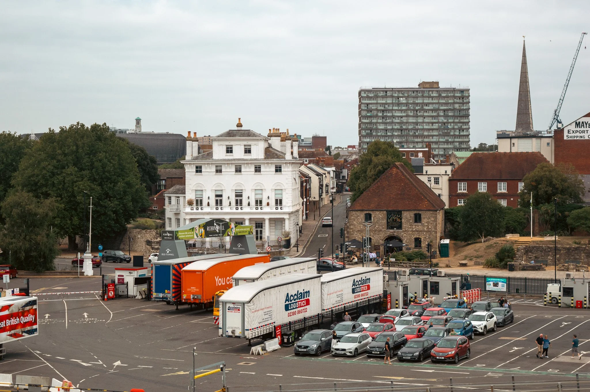 Cityscape featuring a mix of historic and modern buildings, a parking lot filled with cars, trucks, and shipping containers, overcast sky with gray clouds, and some trees.