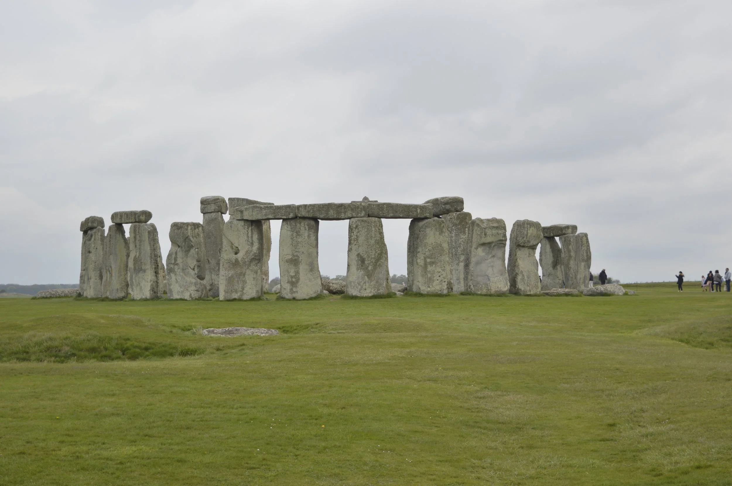 Stonehenge, a prehistoric monument with large standing stones arranged in a circular pattern, is seen on a cloudy day with grass in the foreground and small groups of people walking around.