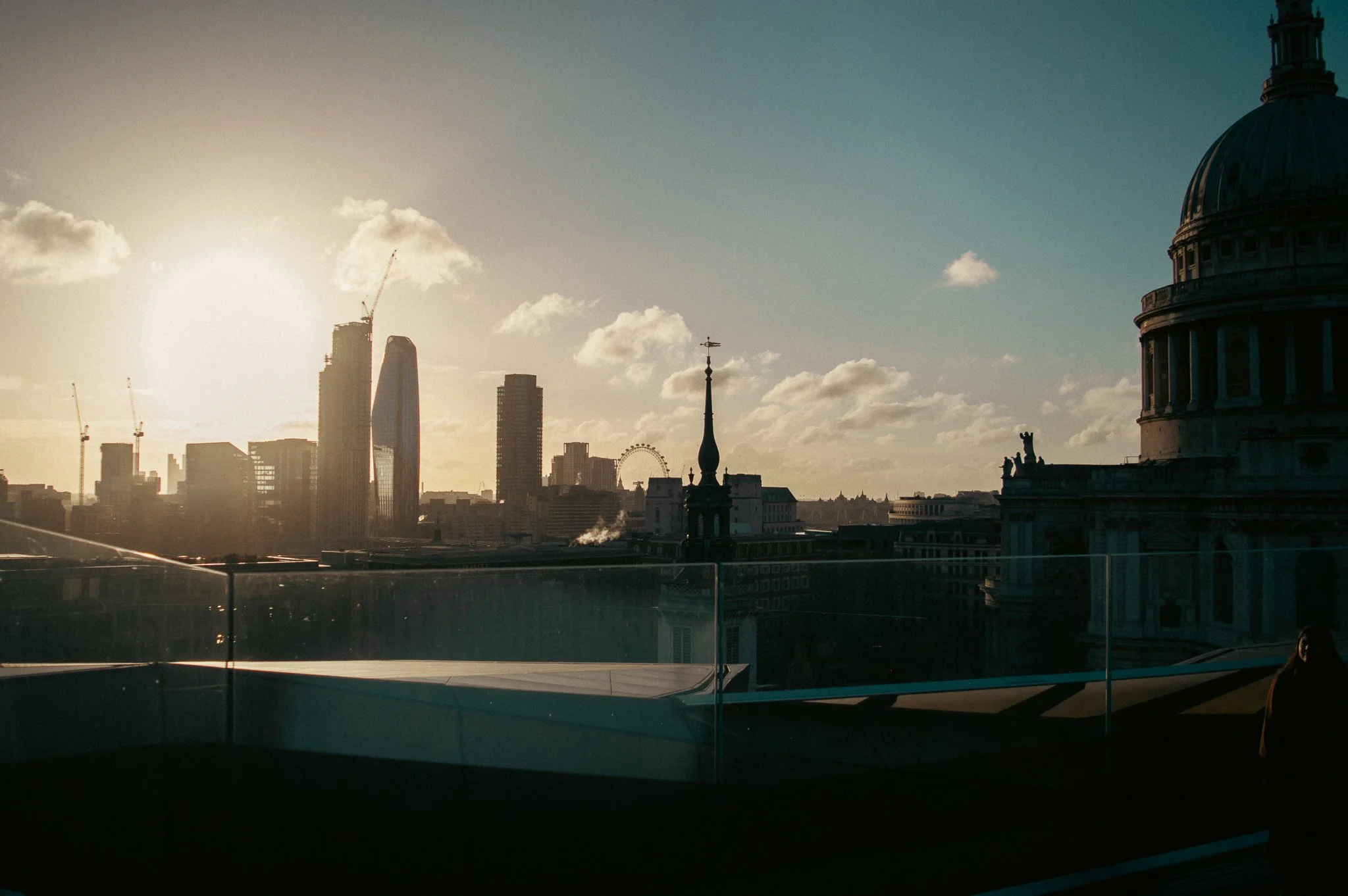 City skyline with modern skyscrapers, a tower with a weather vane, and the dome of a historic building in the foreground, during sunset.