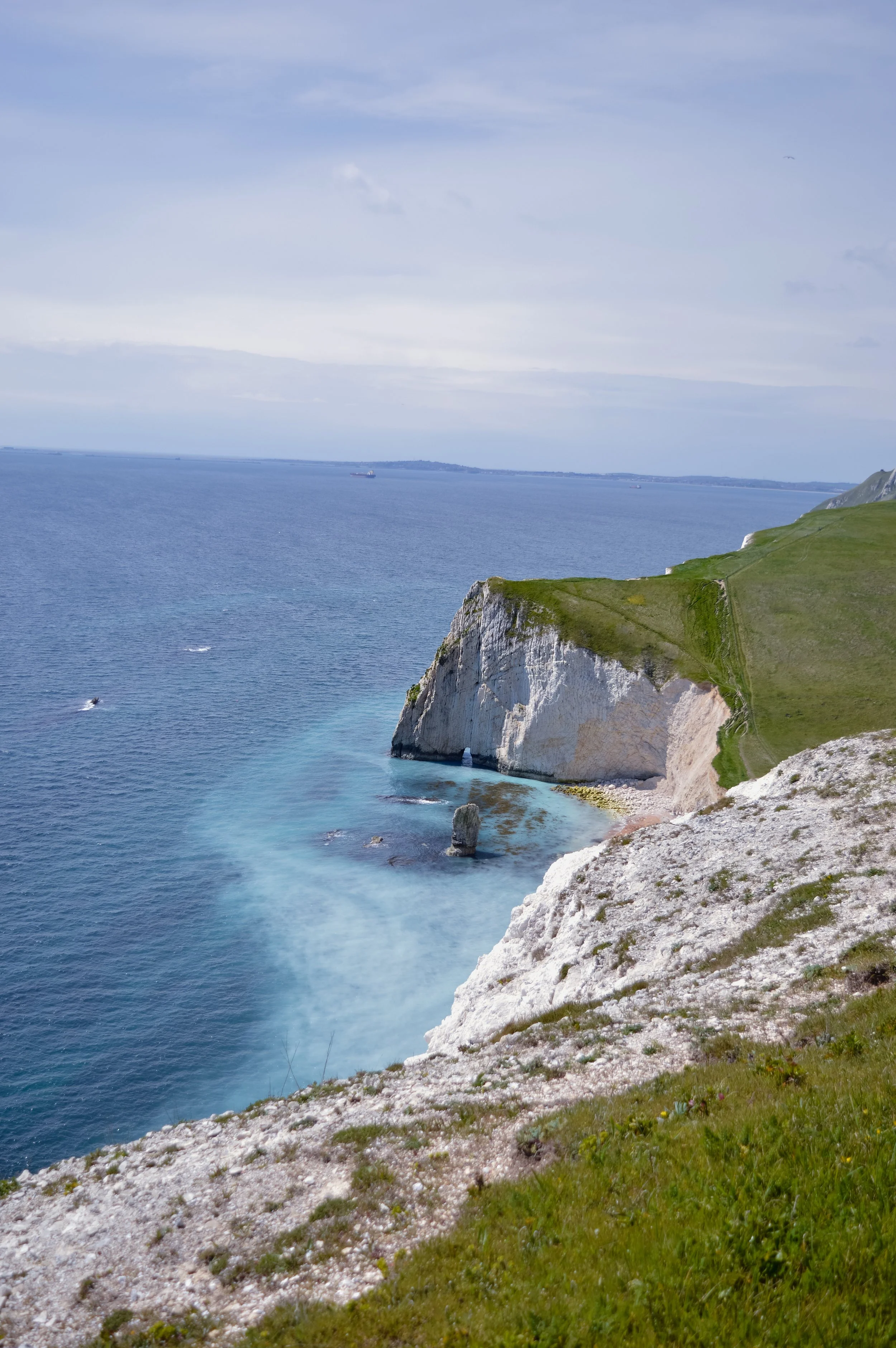 Cliffs overlooking the sea with green grass and rocky slopes, ships in the water, and a cloudy sky above.