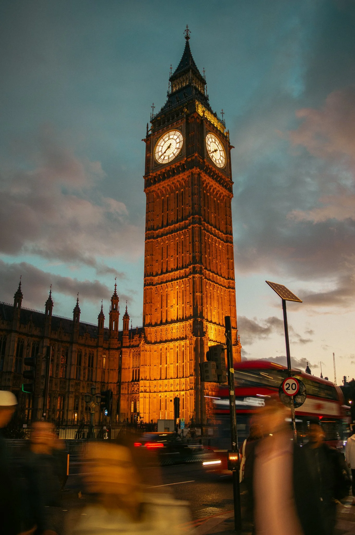 The Big Ben clock tower in London illuminated at dusk with blurred pedestrians and vehicles in motion in the foreground.