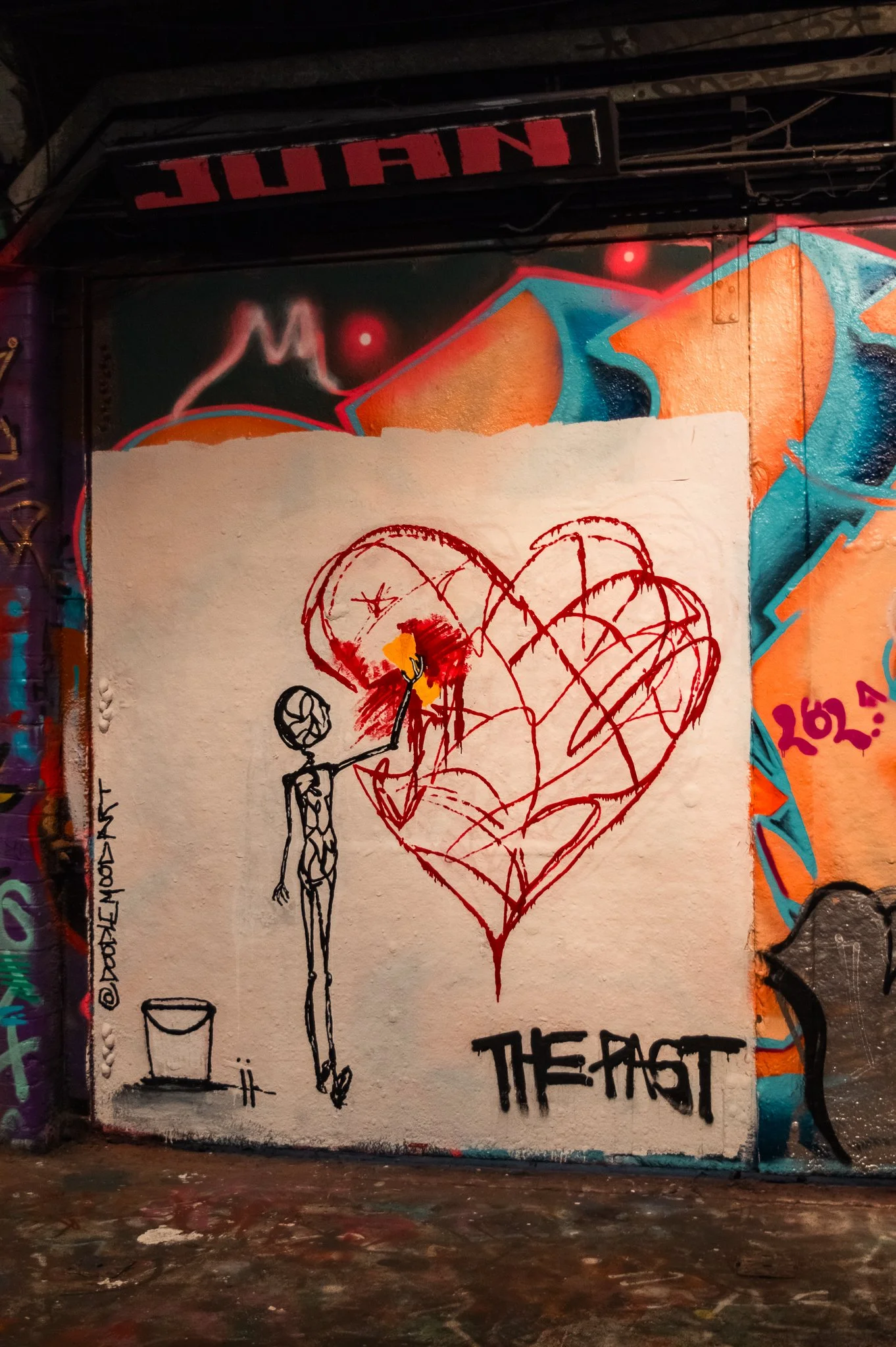 Graffiti art on a wall featuring a stylized figure with a large heart outlined in red and black. The figure, sketched in black, has a yellow patch and stands next to the heart. The background is decorated with colorful abstract shapes and patterns.