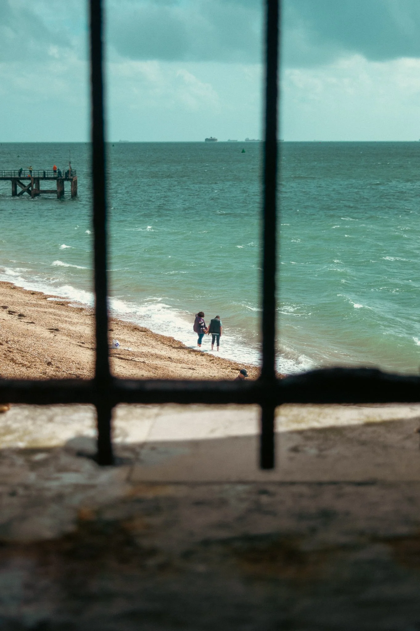 Two people walking along a beach with a pier on the left and ships in the distant ocean, viewed through a window with black bars.