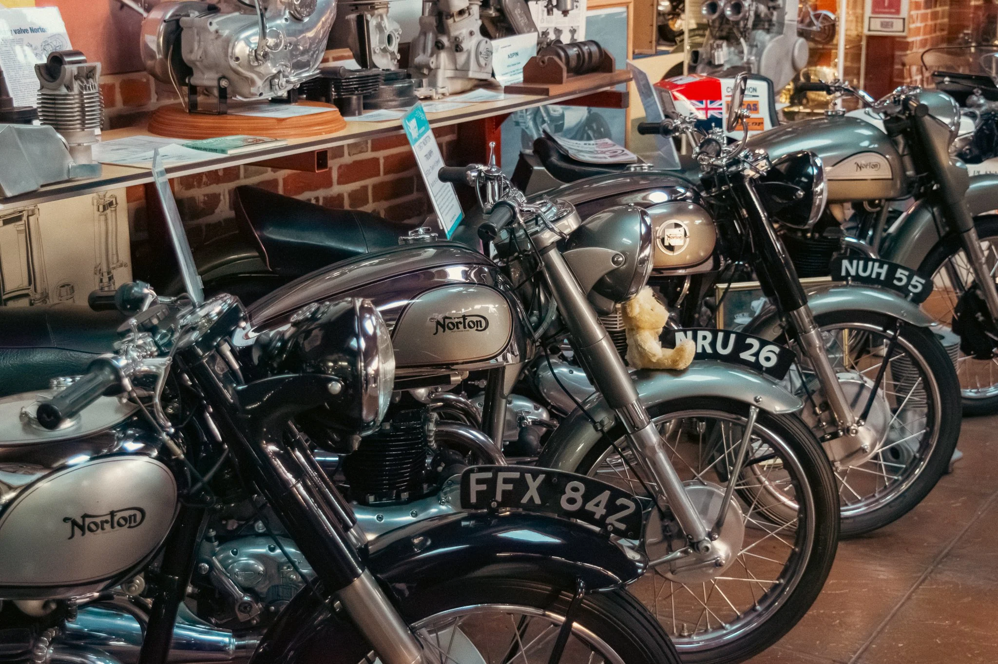 Several vintage Norton motorcycles on display indoors, with multiple engine parts and motorcycle models lined up in front of brick walls.