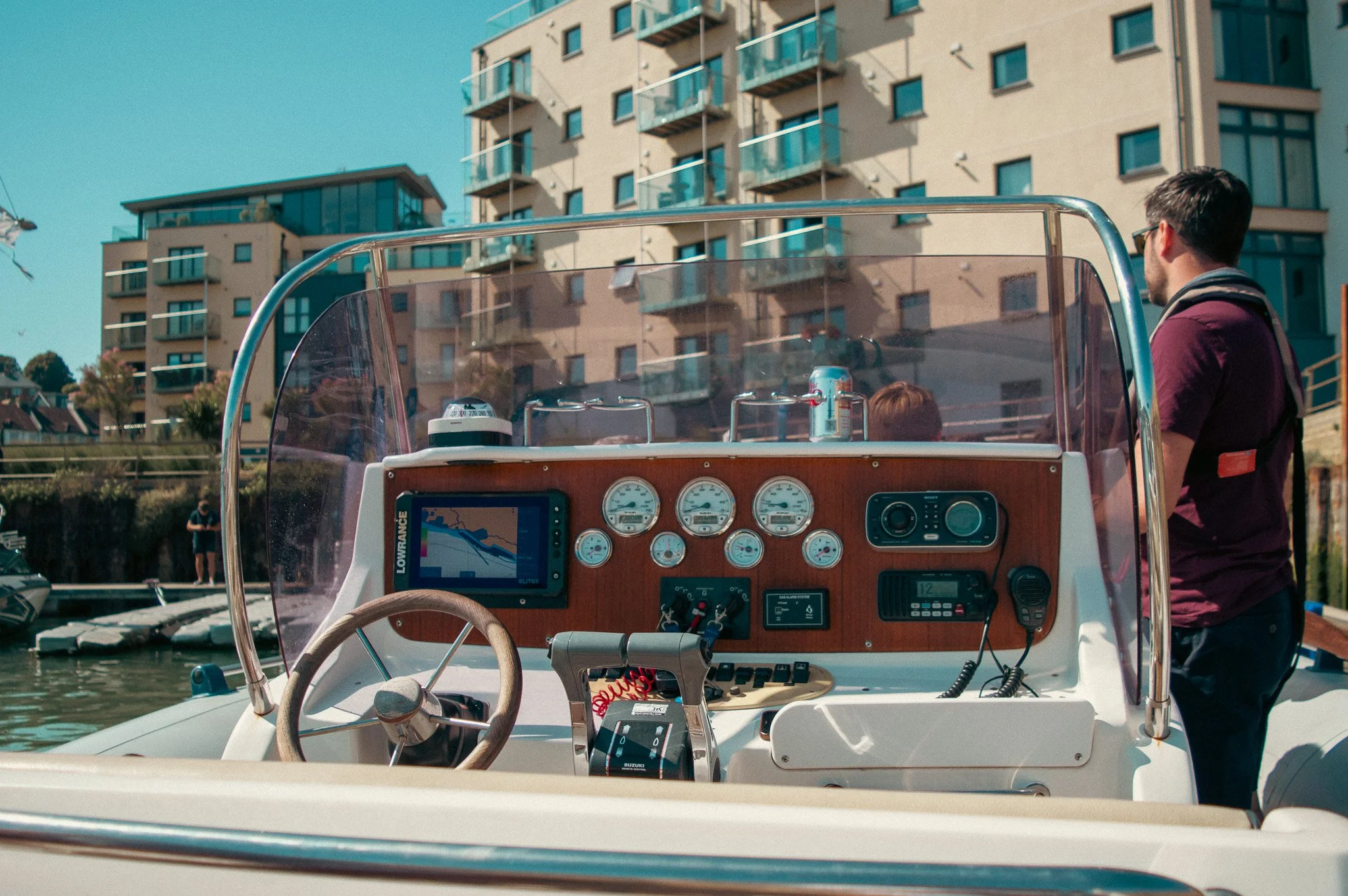 View of the cockpit of a boat with navigational equipment, a man standing on the dock, and residential buildings with balconies in the background.