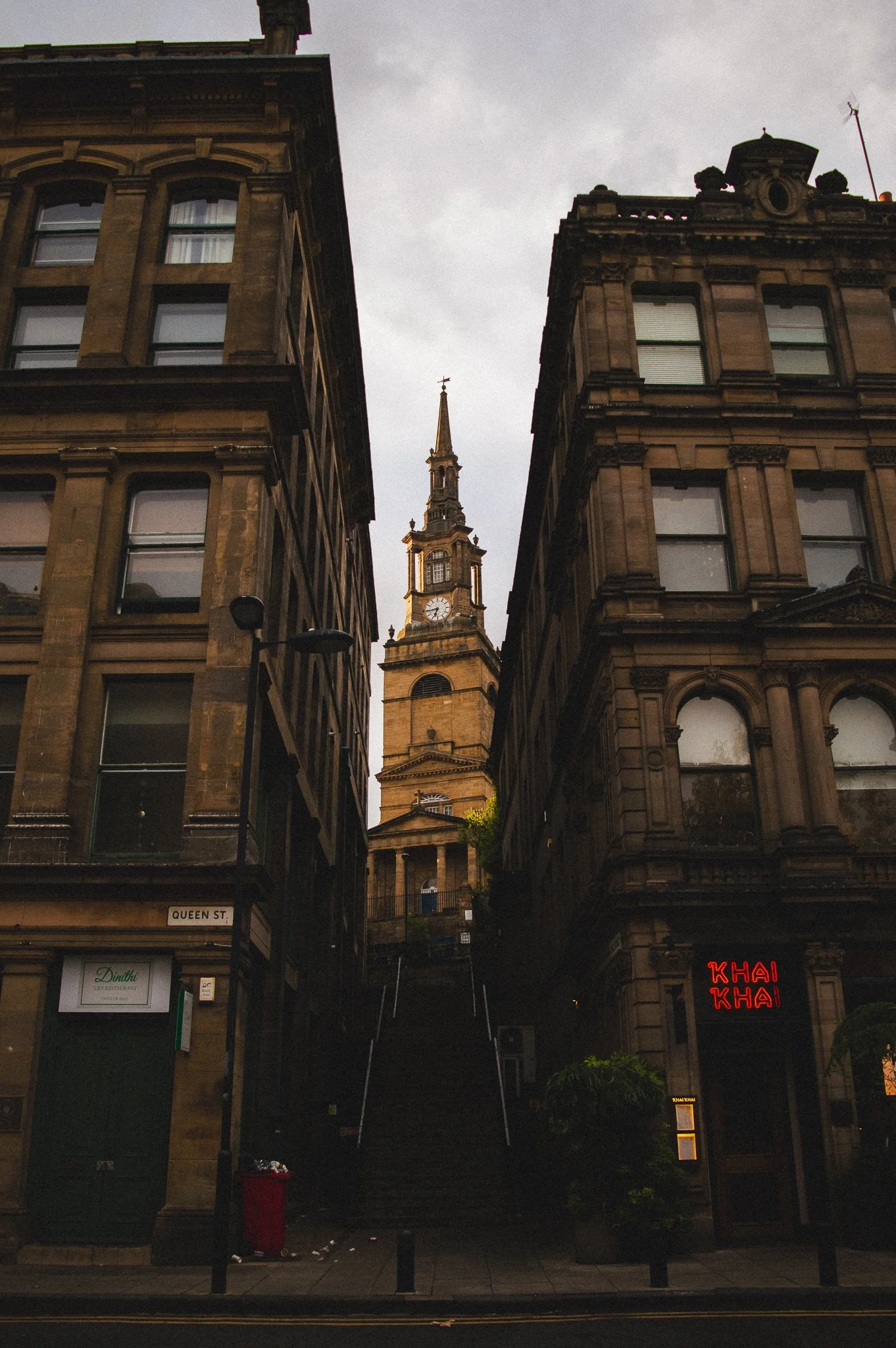 View of historic buildings on Queen Street with a church steeple in the background, a neon sign reading 'Khai Khai', and stairs leading up between the buildings. Overcast sky.