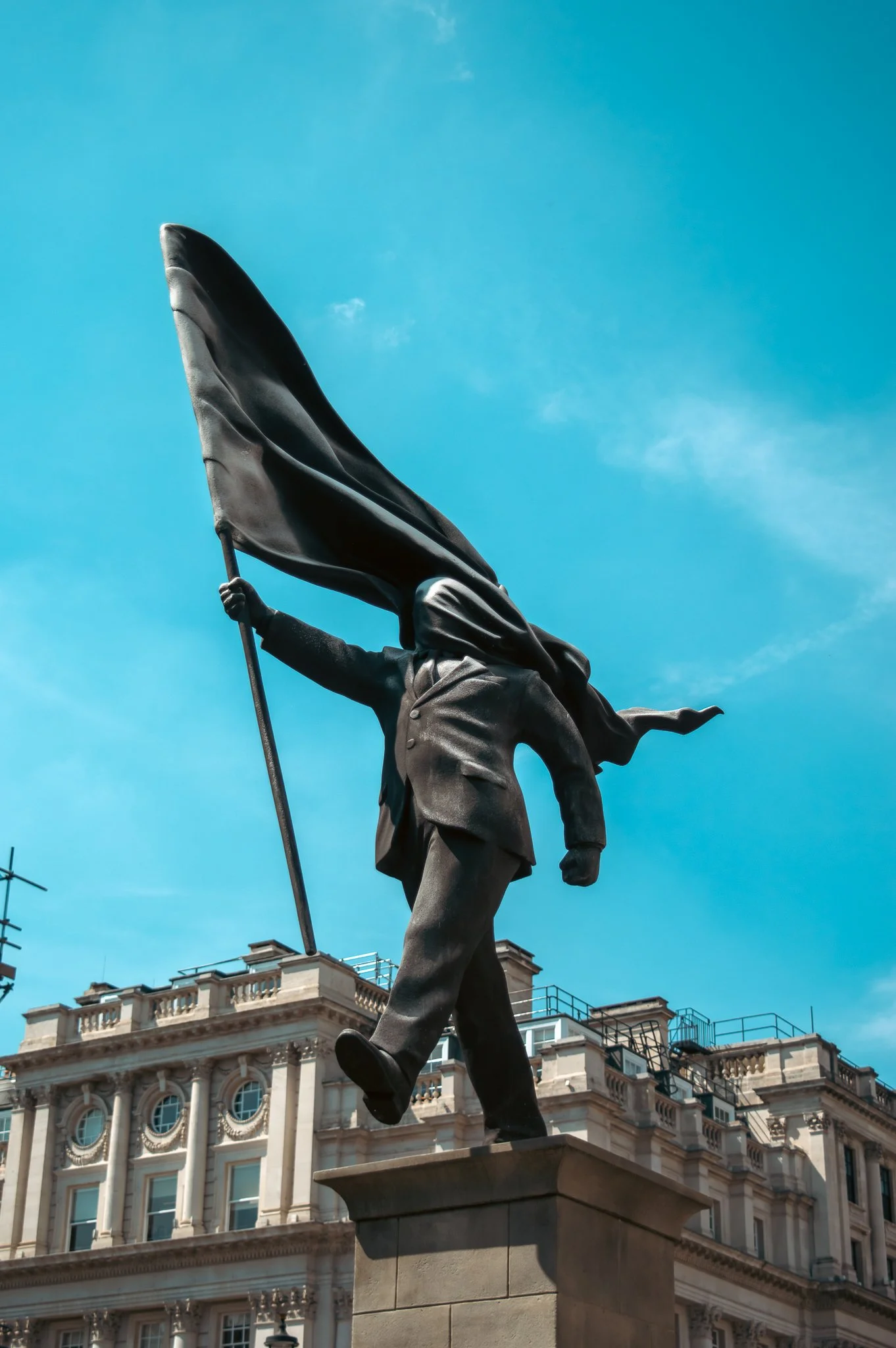 A bronze statue of a man walking with a flag, set against a historic building with ornate architecture, and a bright blue sky.