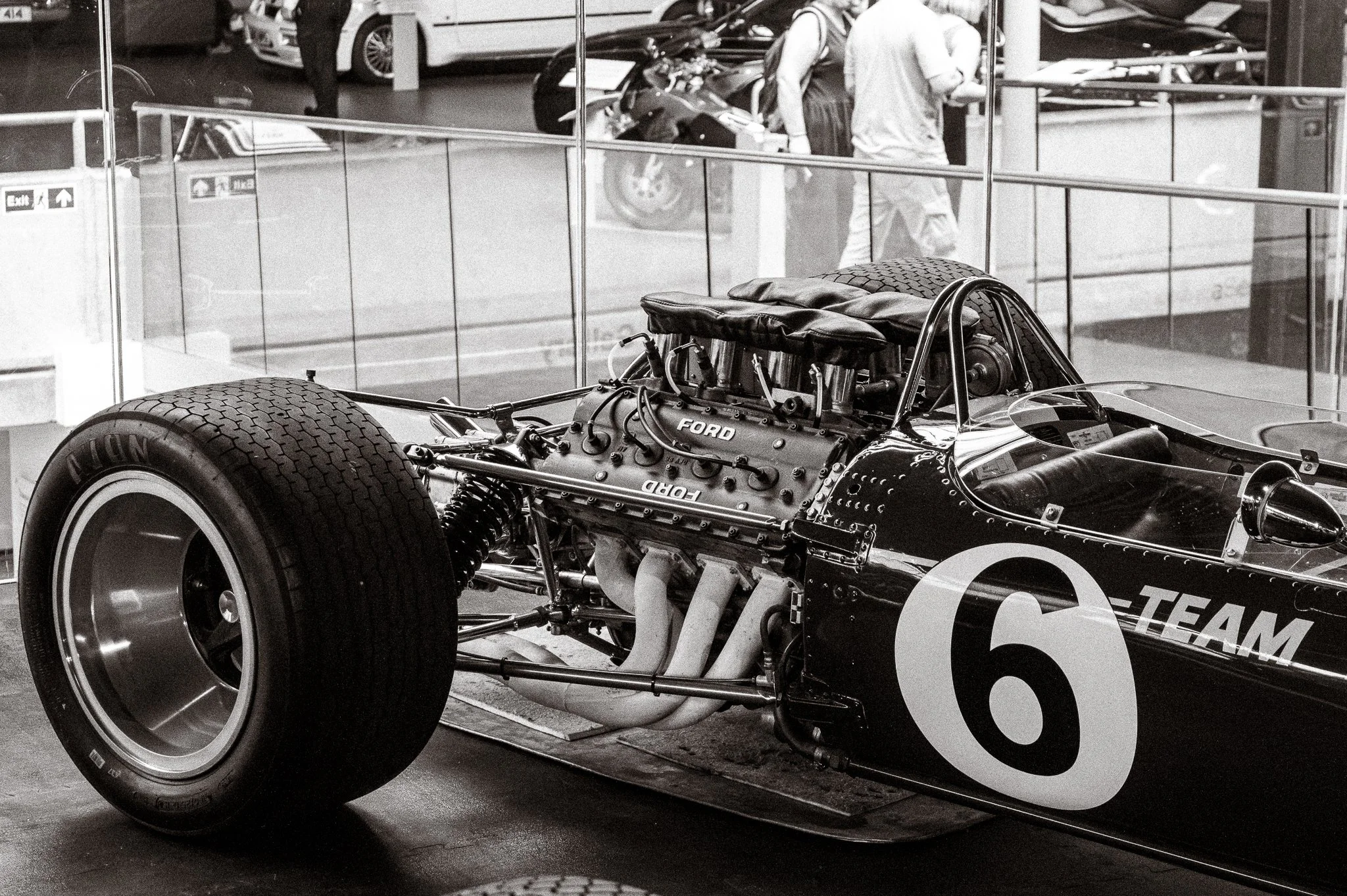A vintage race car with the Ford logo, showing its exposed engine and large tires, displayed in a museum or exhibition space behind a glass barrier, with people and other cars visible in the background.