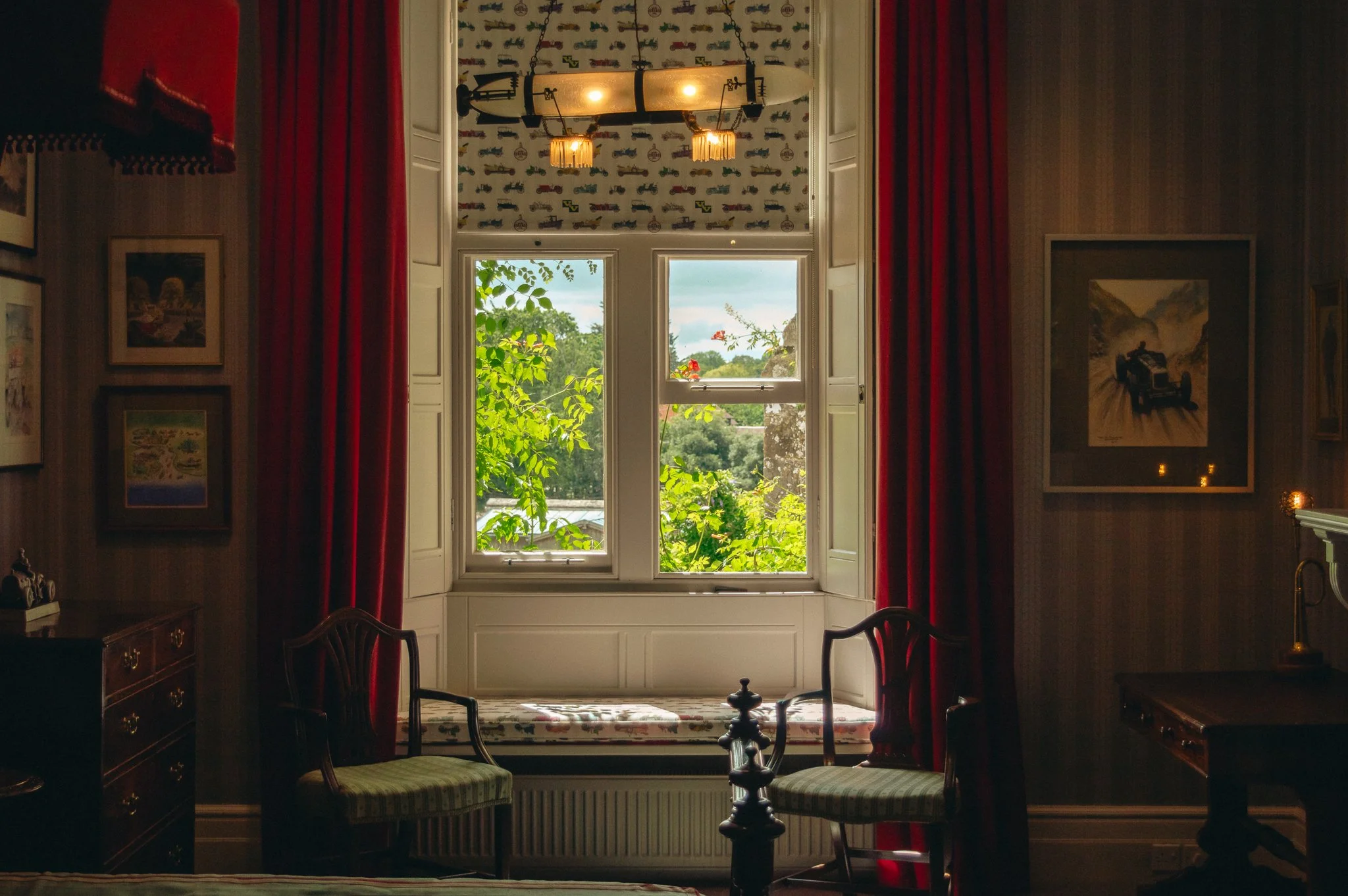 A cozy room with a large window overlooking greenery and a cloudy sky, framed by red curtains. The room has artwork on the walls and a vintage chandelier hanging from the ceiling.