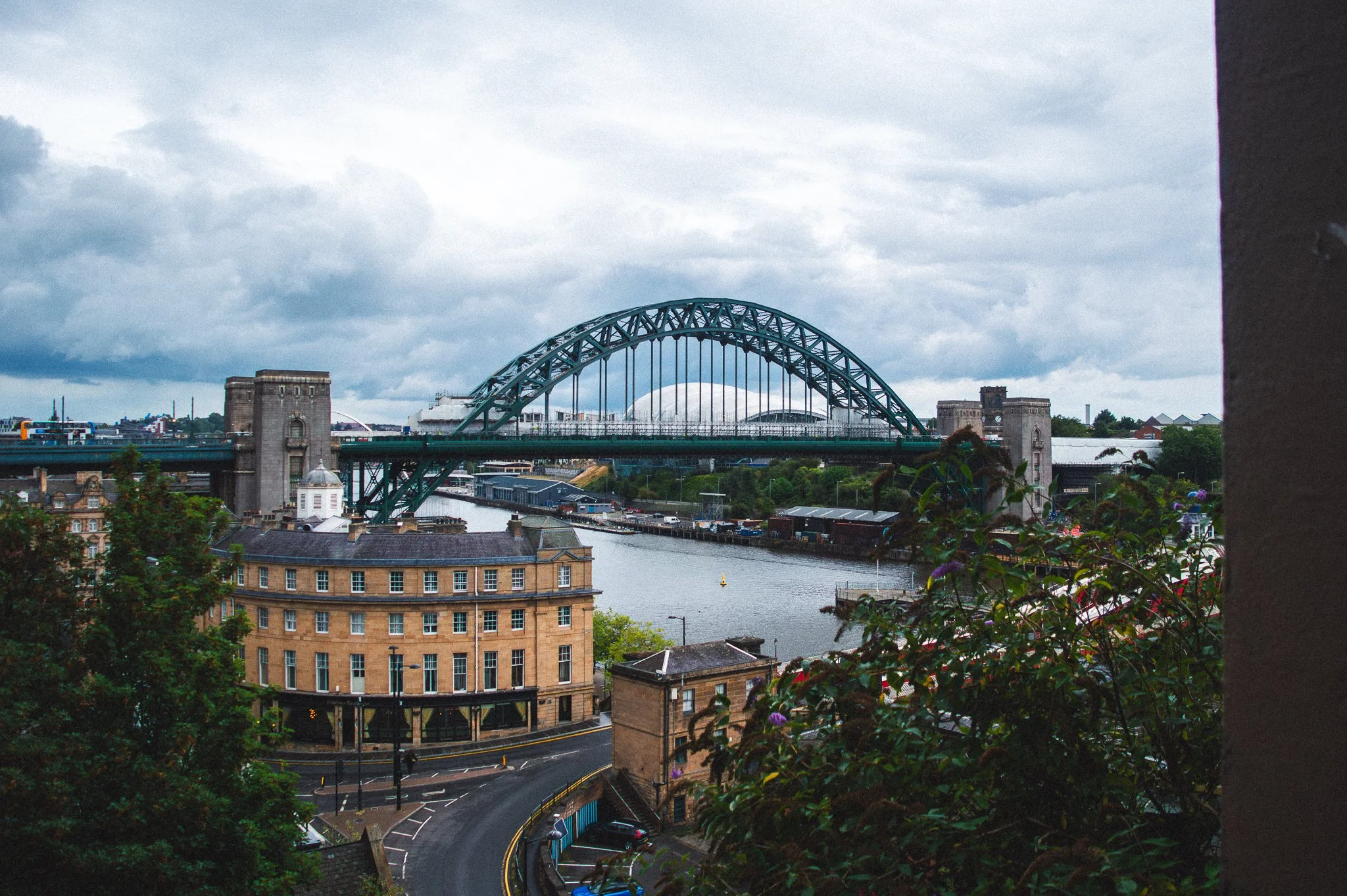 Cityscape featuring a river, a bridge with a large arch, and multiple buildings under a cloudy sky.