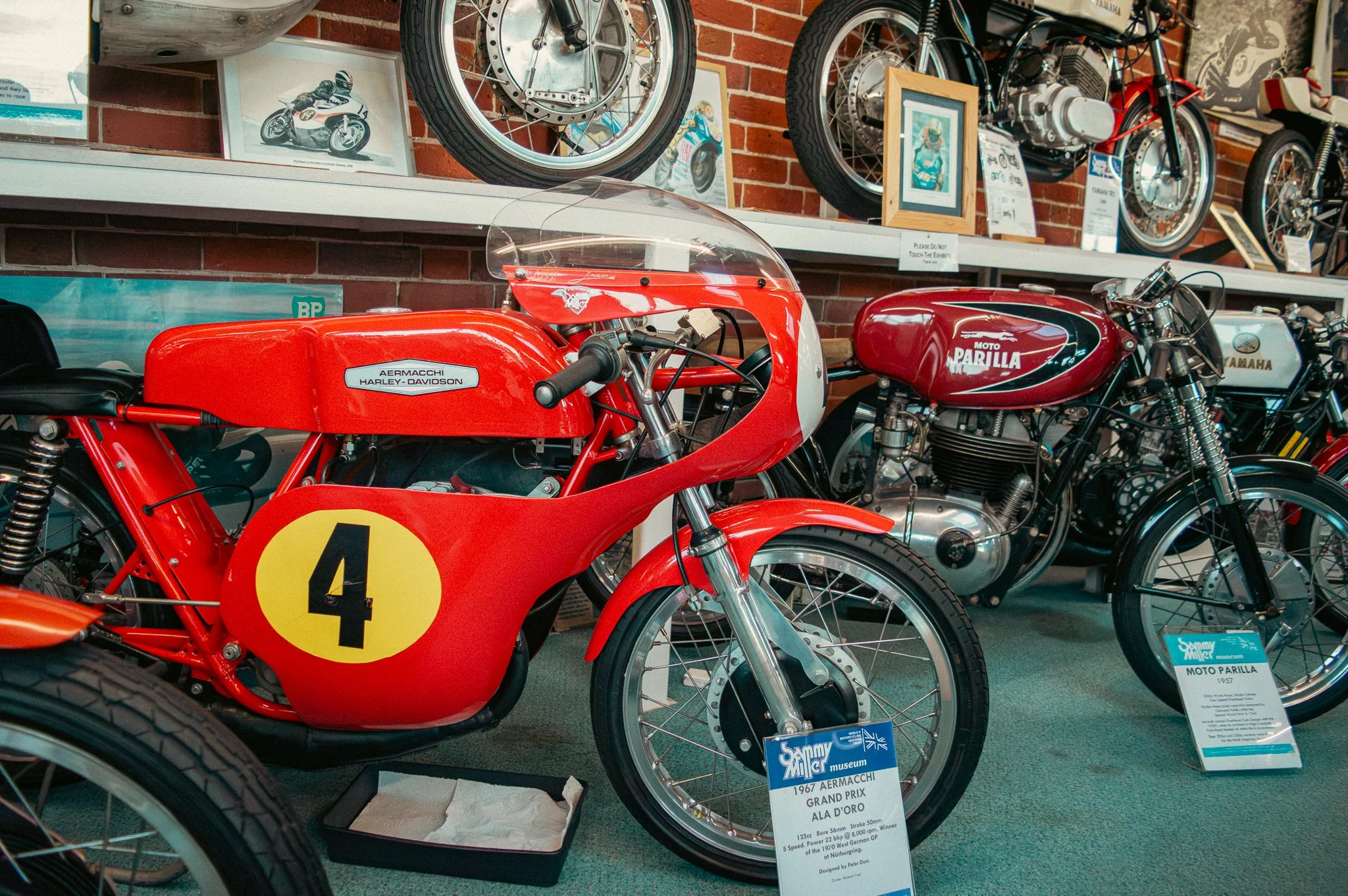 Photo of vintage racing motorcycles on display at a museum, with one prominently showing a red Aermacchi Harley-Davidson with a yellow number 4, and another Yamaha Moto Parilla. Framed pictures of bikes and plaques are on the wall behind them.