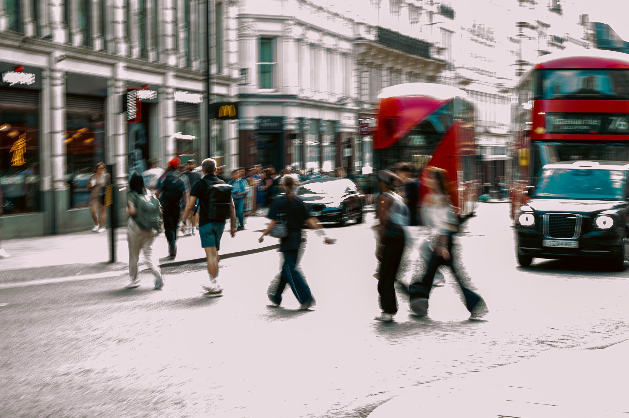 Street scene in a city with pedestrians crossing the street, red double-decker buses, and storefronts on a sunny day.