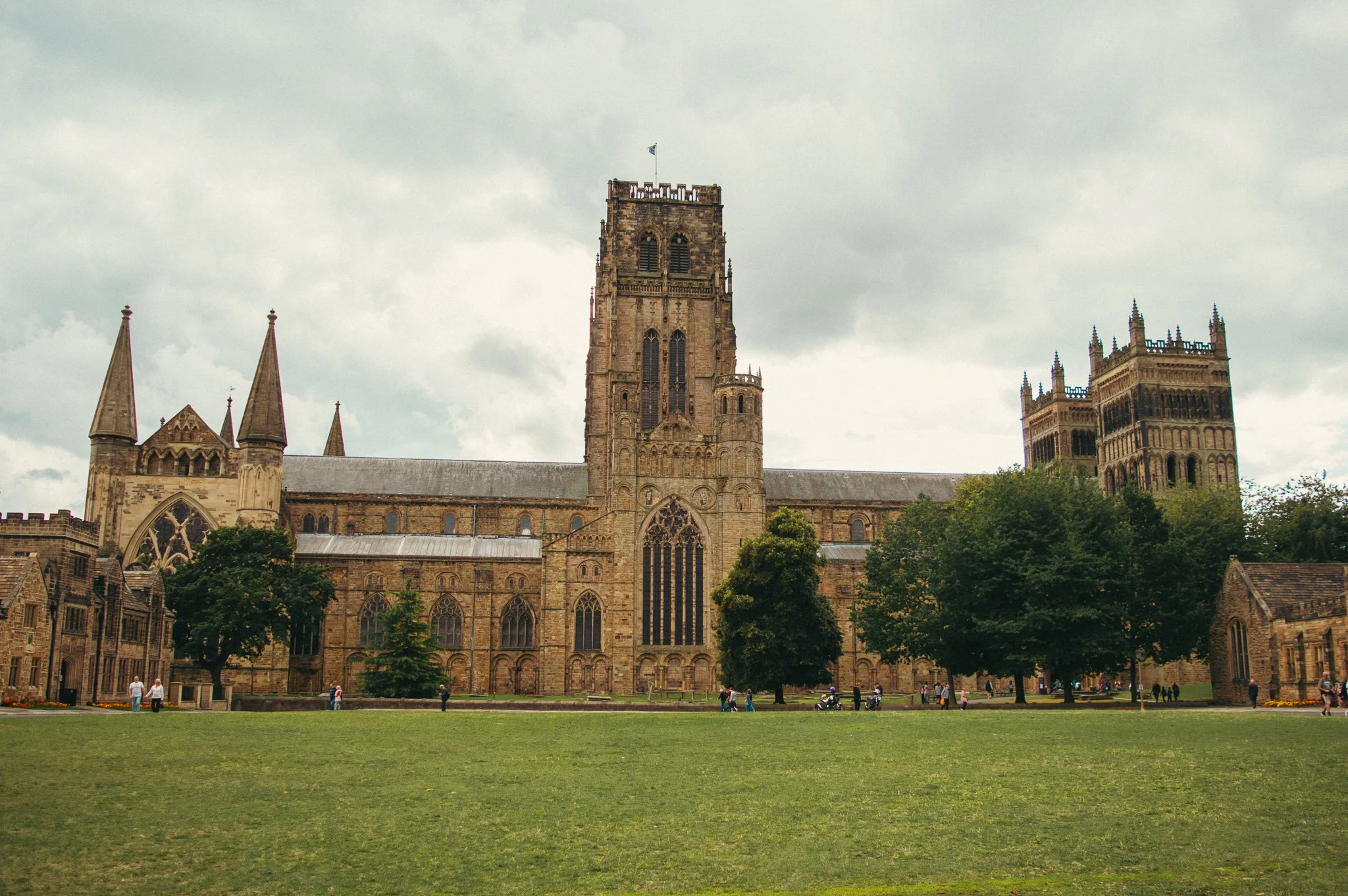A historic stone cathedral with tall towers and intricate architecture, set in a grassy yard with trees, people, and cloudy sky.