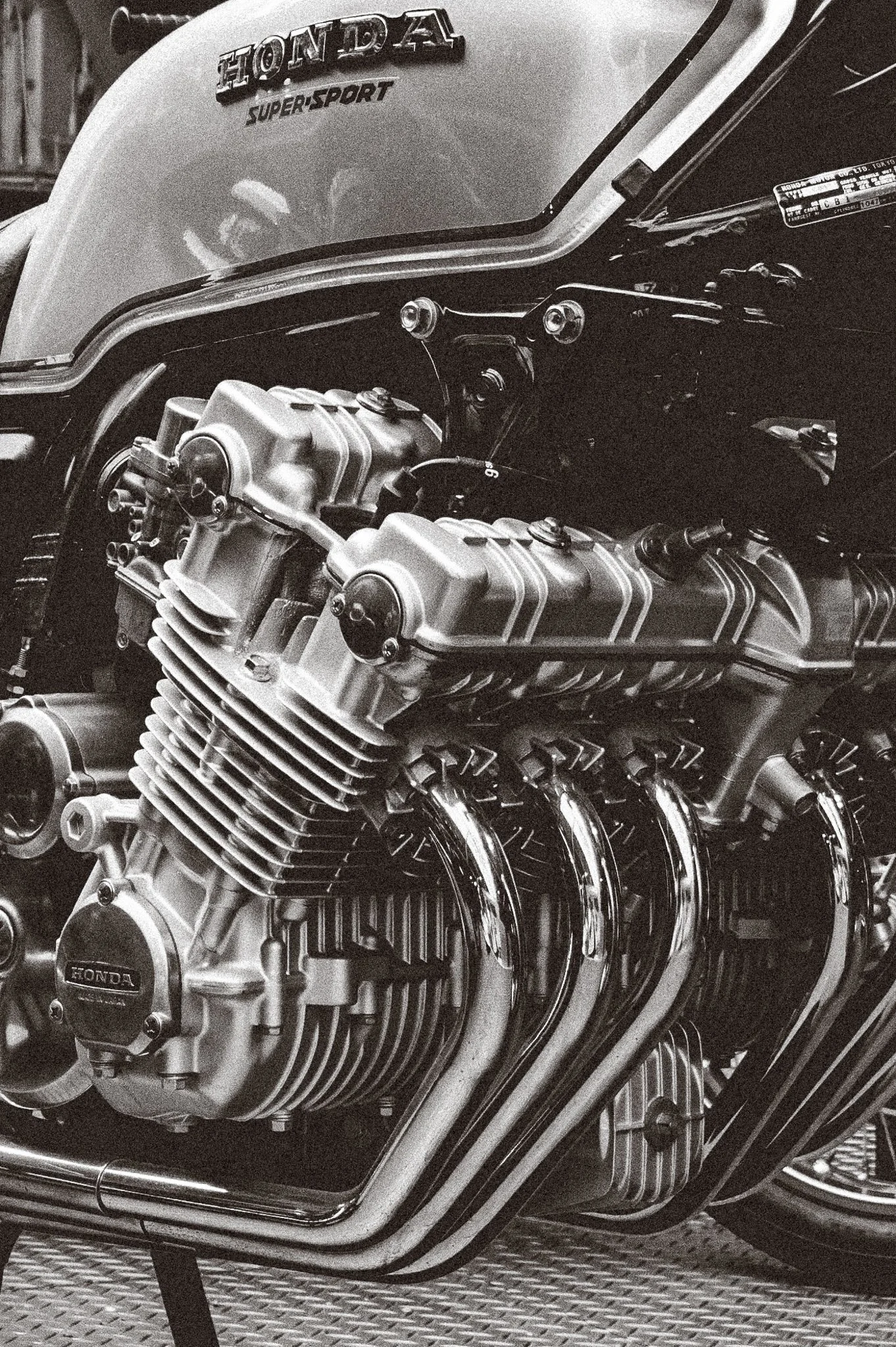 A close-up black and white photo of a Honda motorcycle engine with visible chrome pipes and metallic components, with the 'Honda' logo on the engine casing, and the motorcycle's fuel tank with a Honda Super Sport badge.