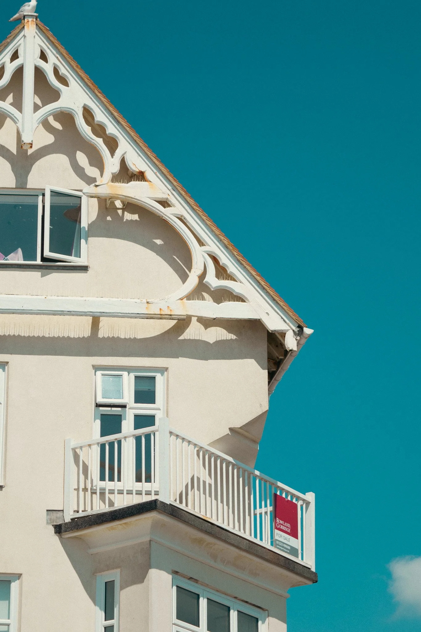 A close-up view of a white building with ornate architectural features, including a balcony with a white railing and multiple windows, under a clear blue sky. There is a red 'For Sale' sign on the balcony railing.