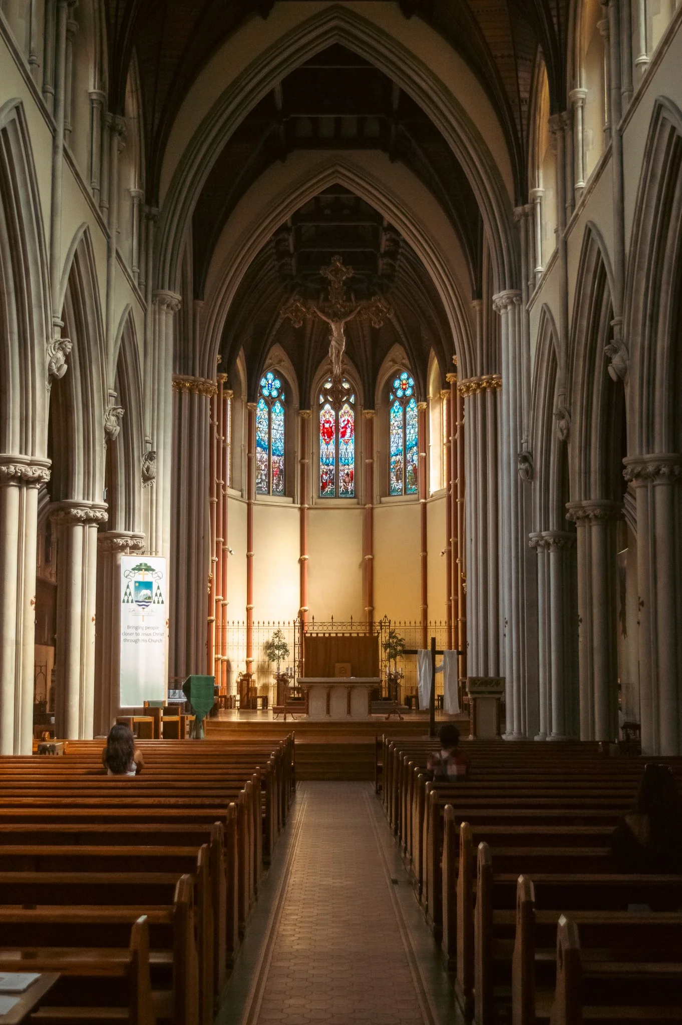 Inside of a church with wooden pews, stained glass windows, and a crucifix hanging above the altar. Two children are seated in the pews.