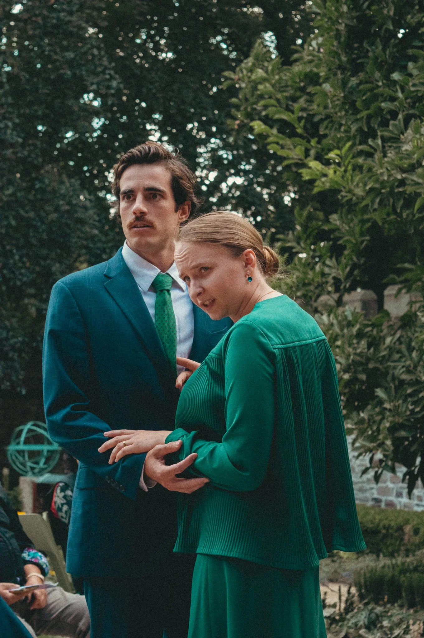 A man in a blue suit with a green tie and a woman in a green dress and blazer holding hands outdoors. They appear to be at a formal event, with trees and other people in the background.