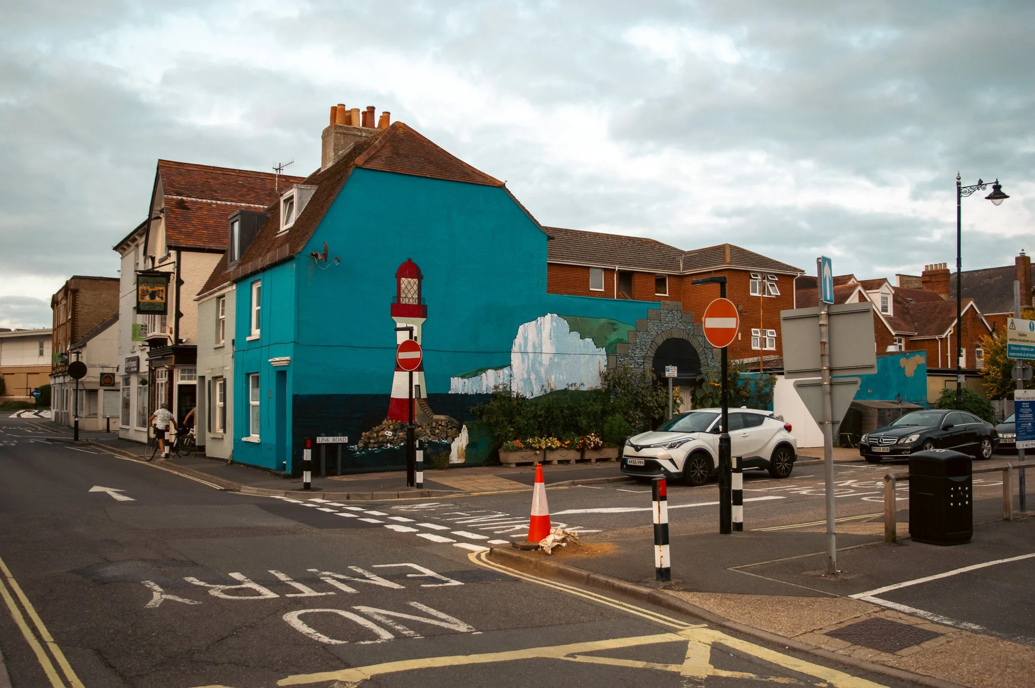 A street scene featuring a mural of a lighthouse on the side of a bright blue building, with cars parked and pedestrians walking nearby under a cloudy sky.