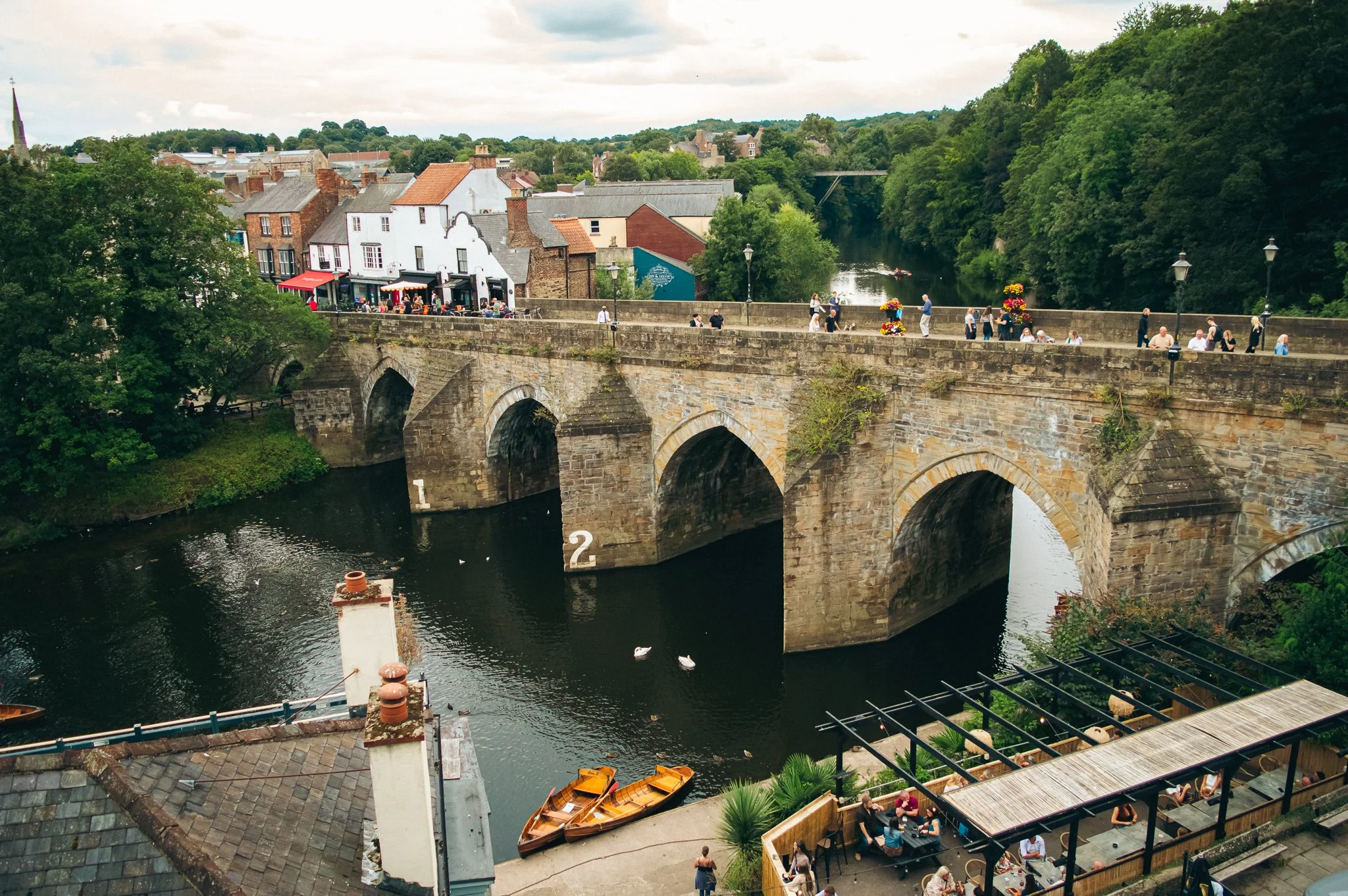 A scenic view of a stone bridge over a river, with people walking along the bridge. There are colorful boats docked by the riverbank and a row of businesses and shops with outdoor seating along the waterfront. Lush green trees line the riverbanks and