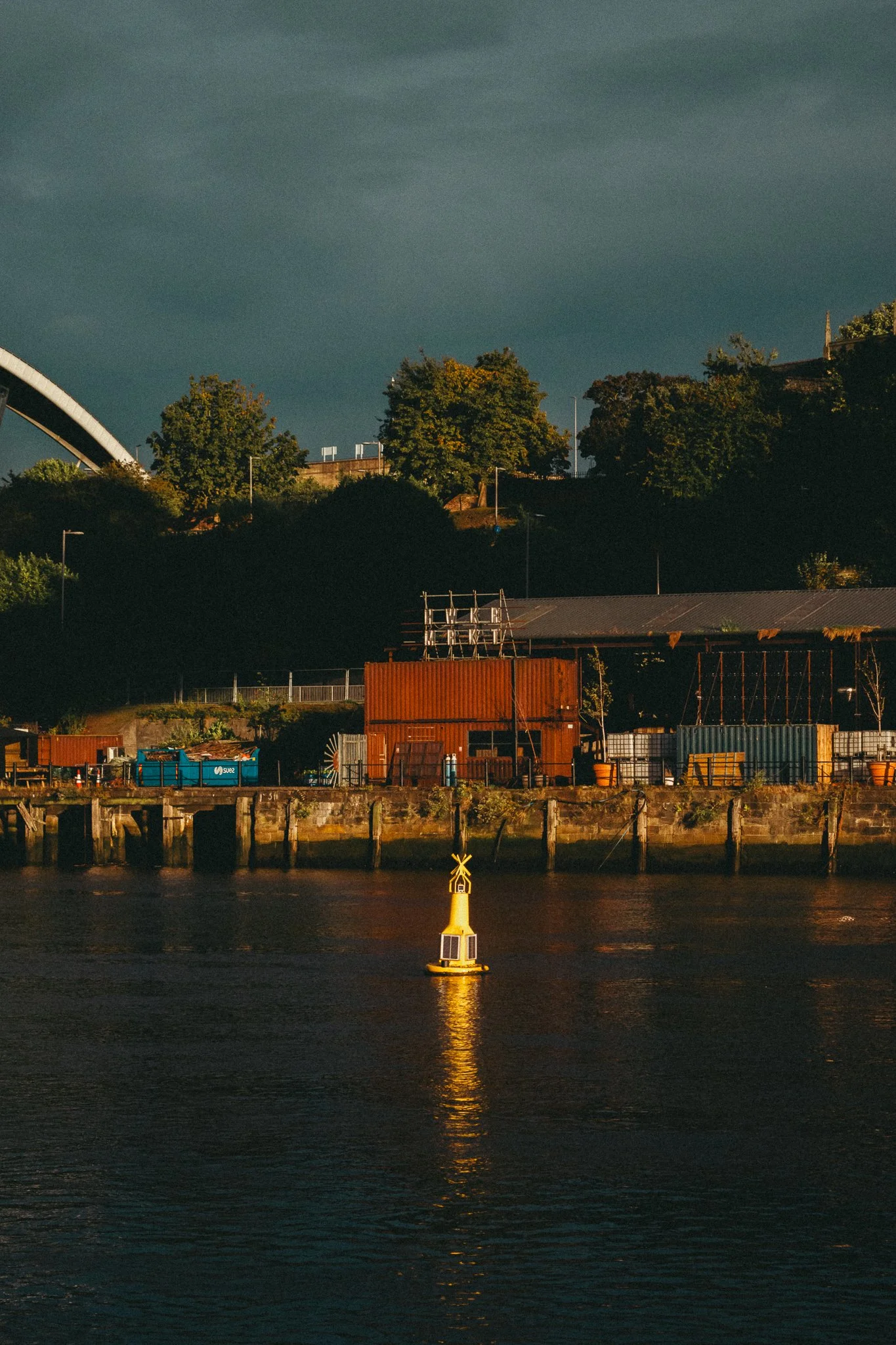 Nighttime scene of a river with a yellow navigation buoy in the foreground, reflecting lights on the water, and a hillside with trees and buildings in the background.