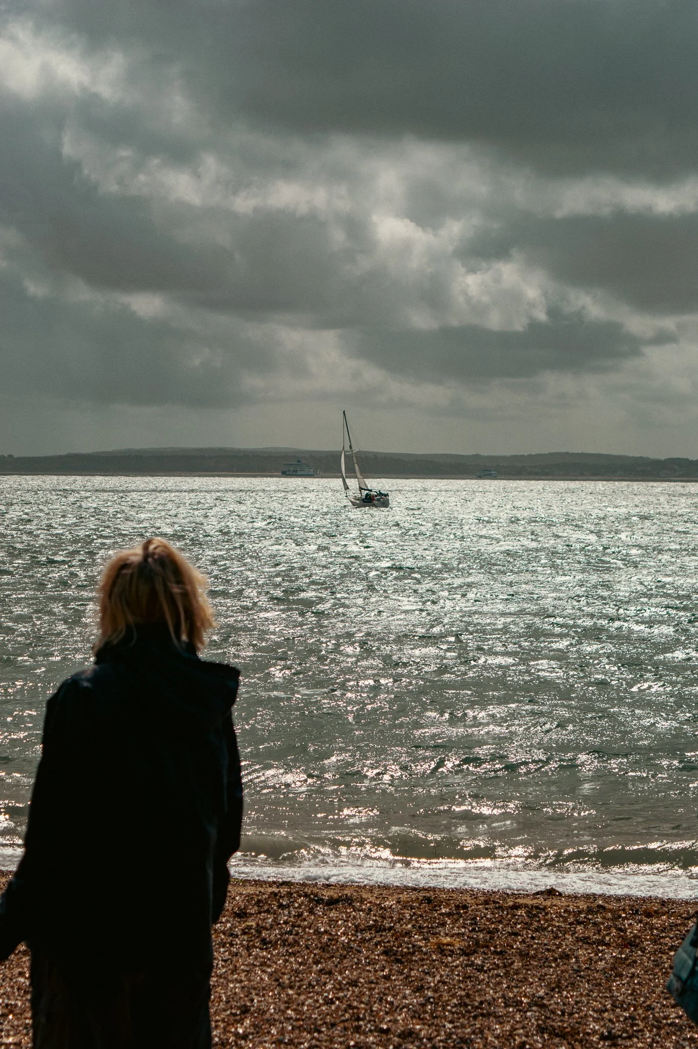 Person with blonde hair wearing a black jacket stands on a pebble beach facing the water with a sailboat and cloudy sky in the distance.