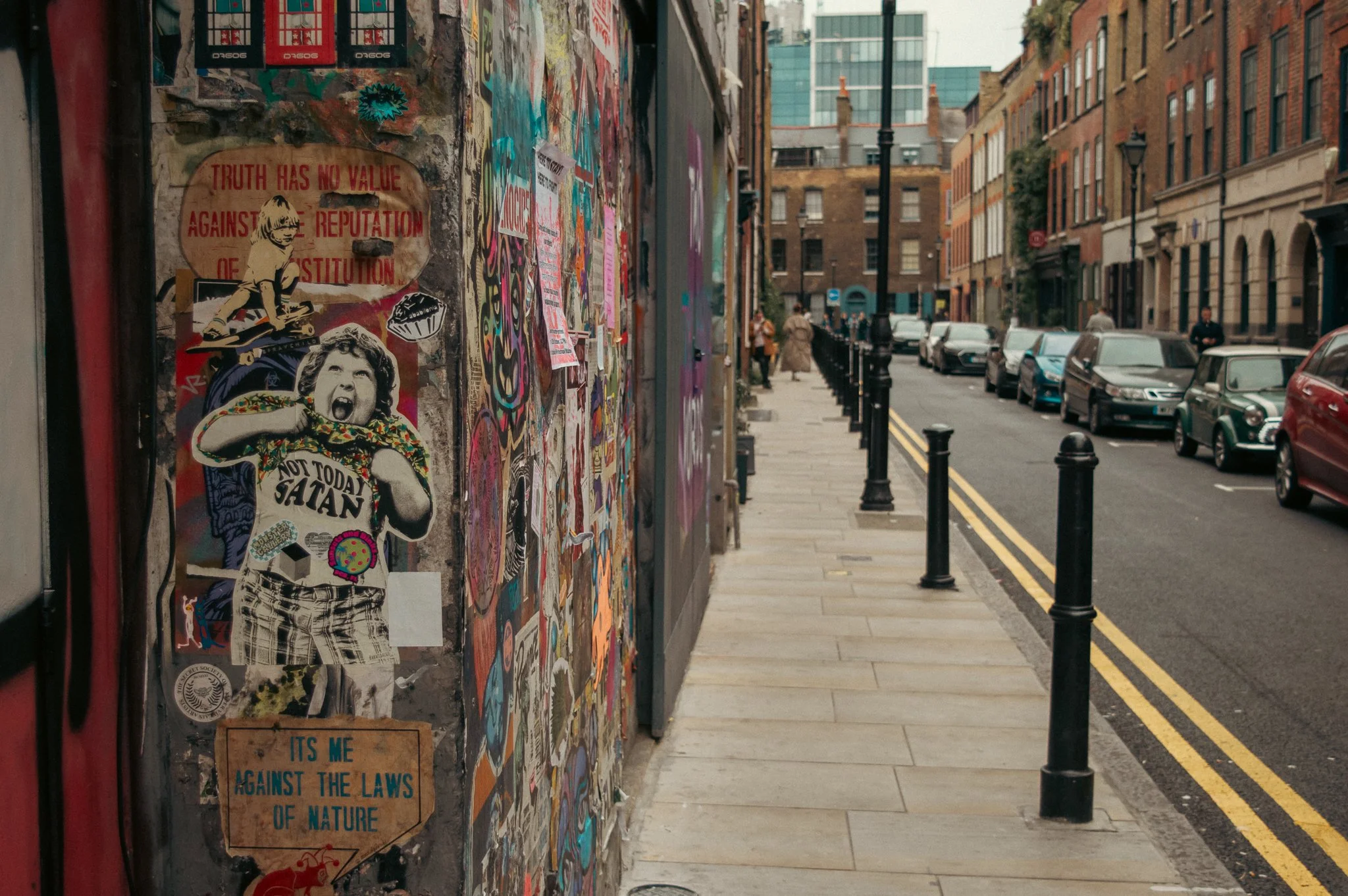 A graffiti-covered wall with posters and stickers in an urban street scene, with parked cars and people walking along the sidewalk.