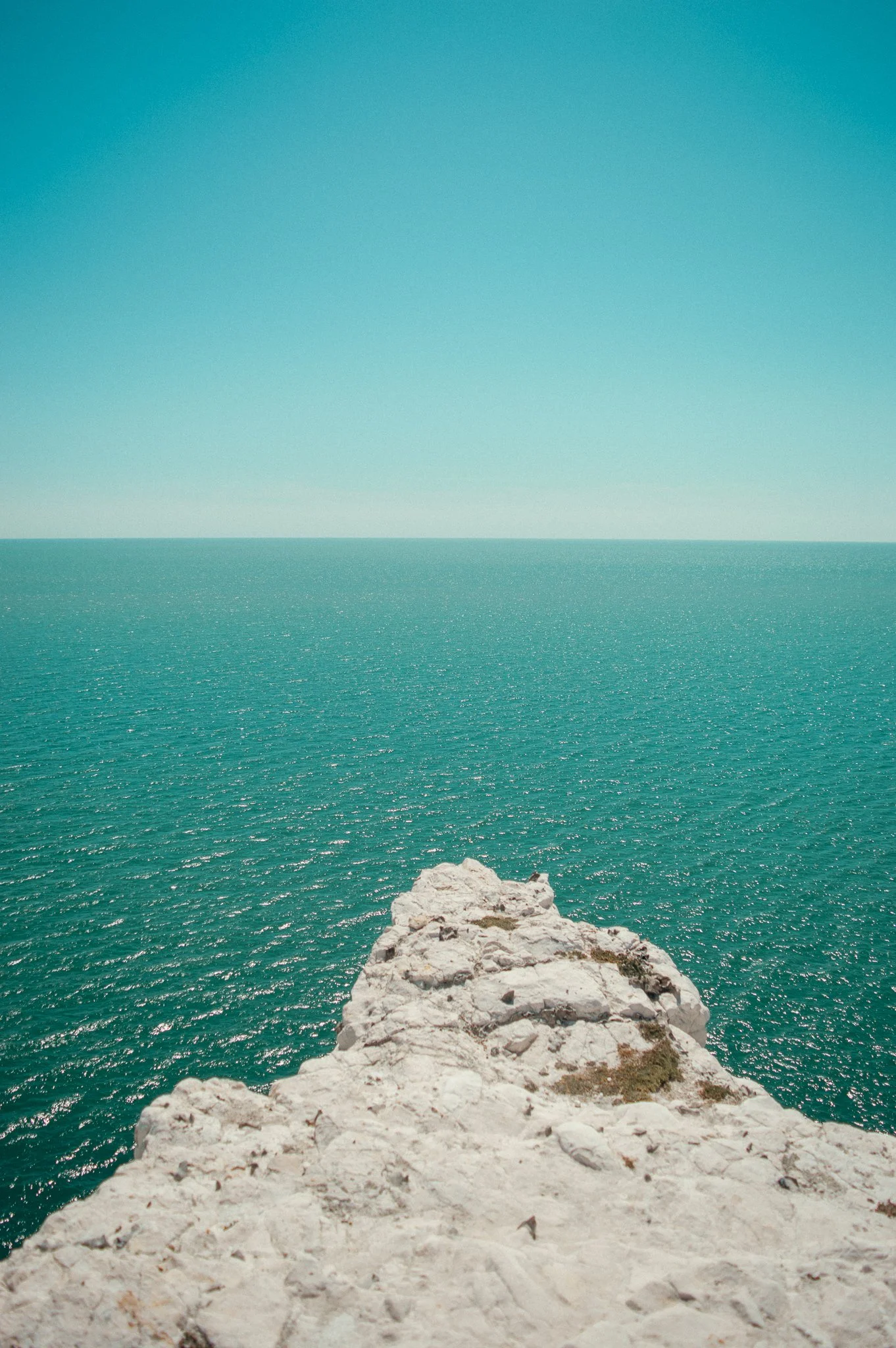 A view from a white rocky cliff overlooking a vast, calm ocean under a clear blue sky.