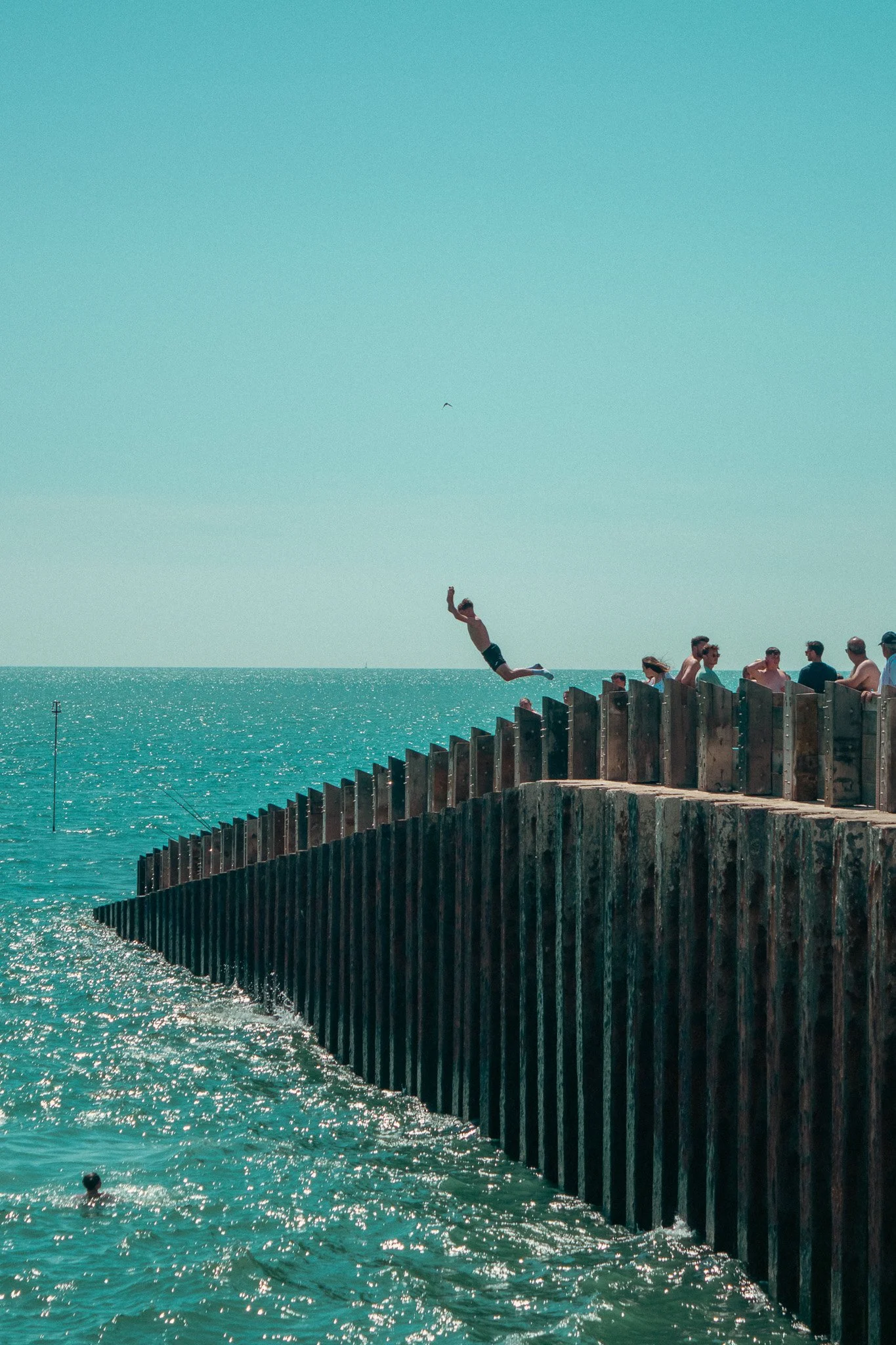 A man jumping from a wooden pier into the ocean with several people sitting on the pier against a bright blue sky.