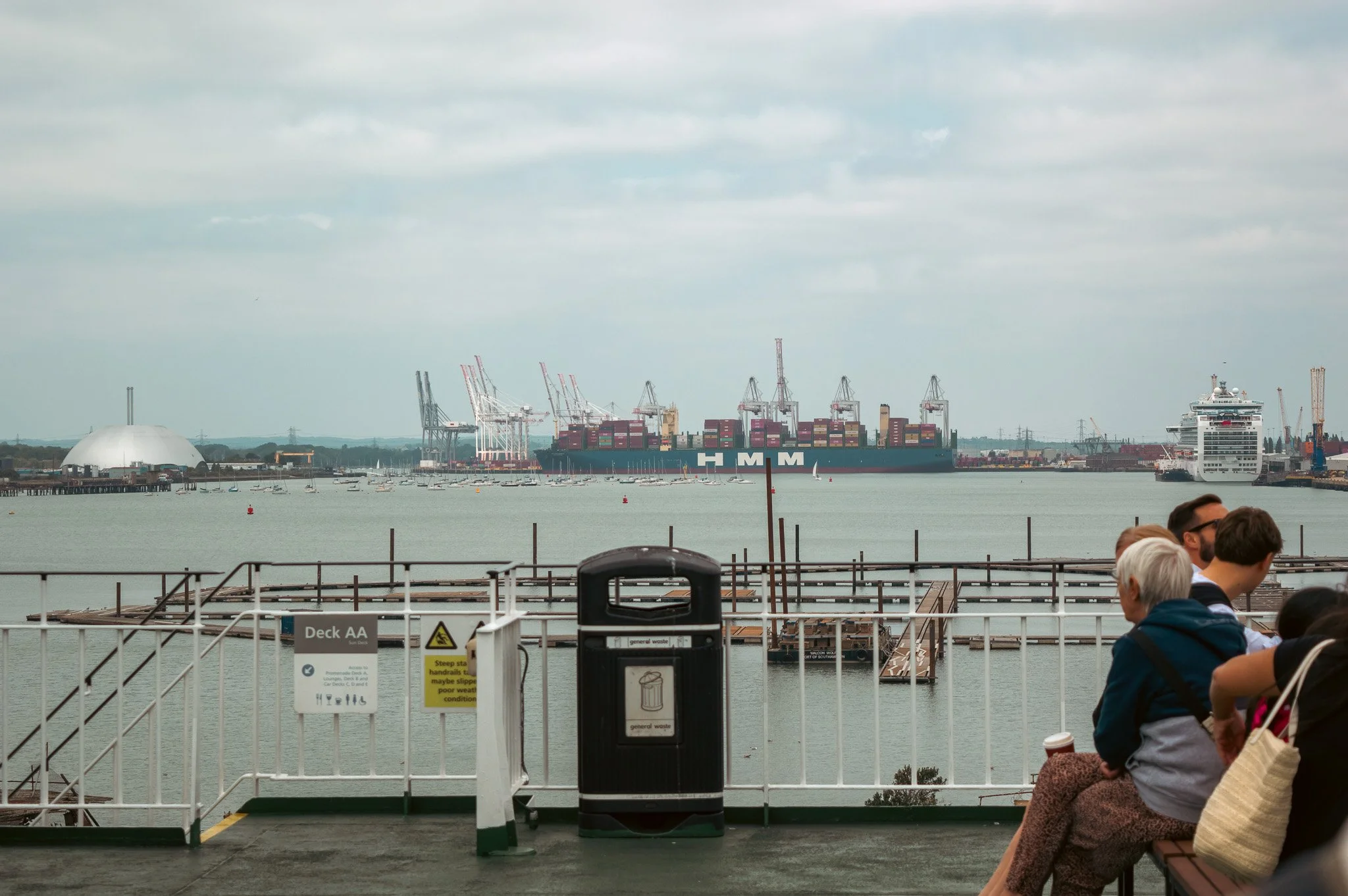 View of a harbor with a large container ship, smaller boats, and port cranes, as well as people sitting on benches in the foreground.