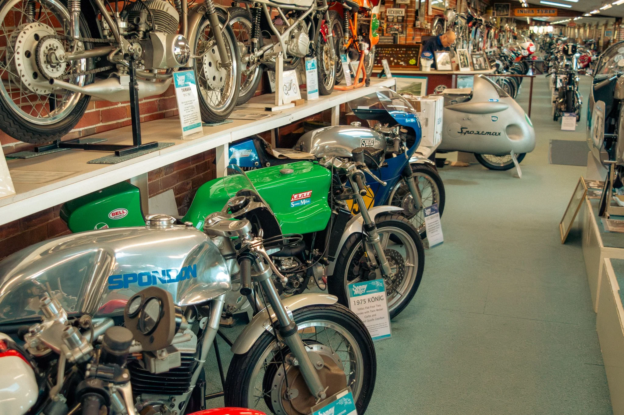 Interior of a museum showcasing vintage motorcycles, including a green racing bike and a silver-bodied motorcycle, with informational signs and background displays of framed pictures and other memorabilia.