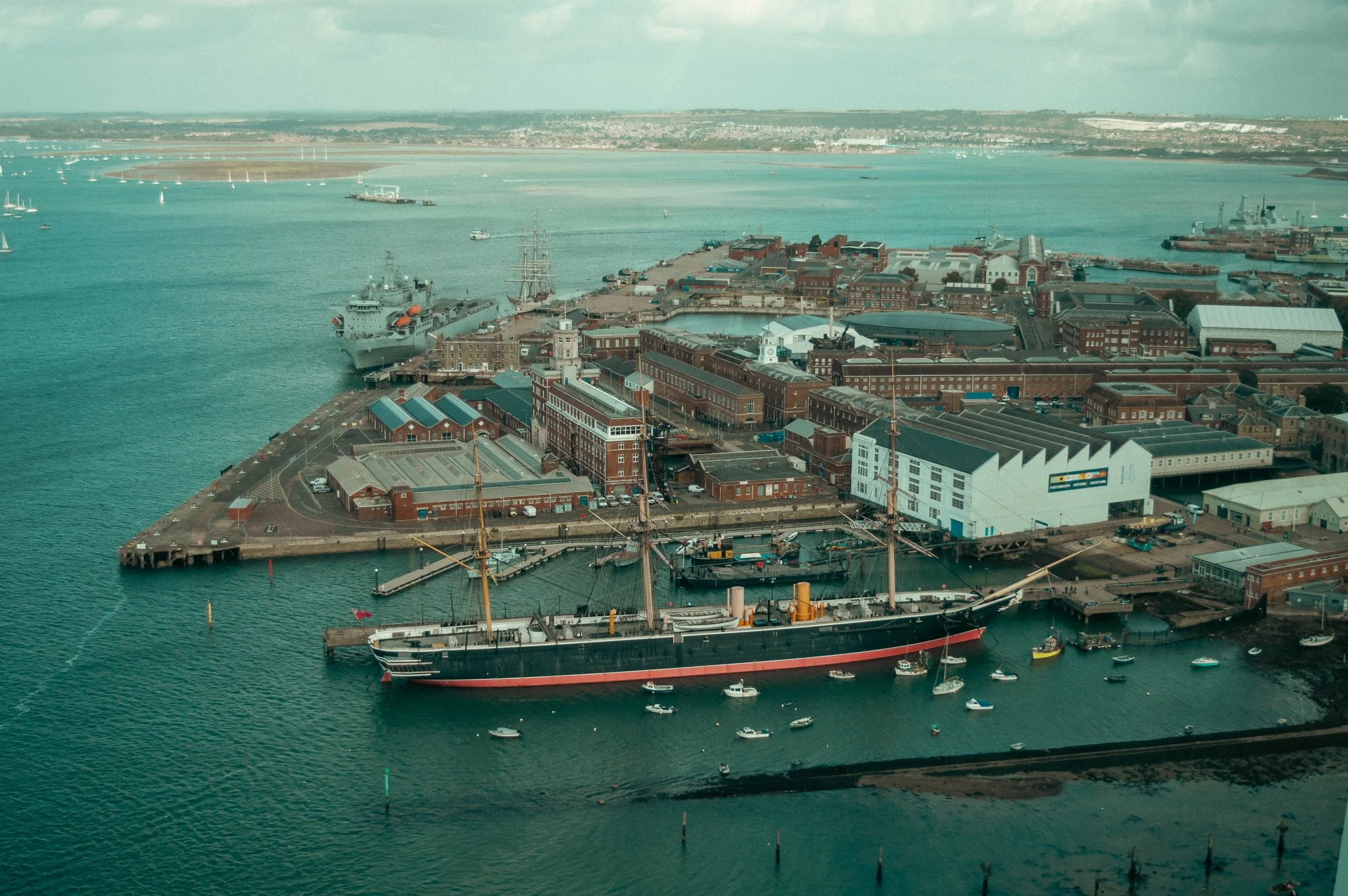 Aerial view of a harbor with ships, sailboats, and a breakwater, featuring historic red brick buildings along the waterfront and a large ship docked at the pier. The background shows a river, distant land, and cloudy sky.