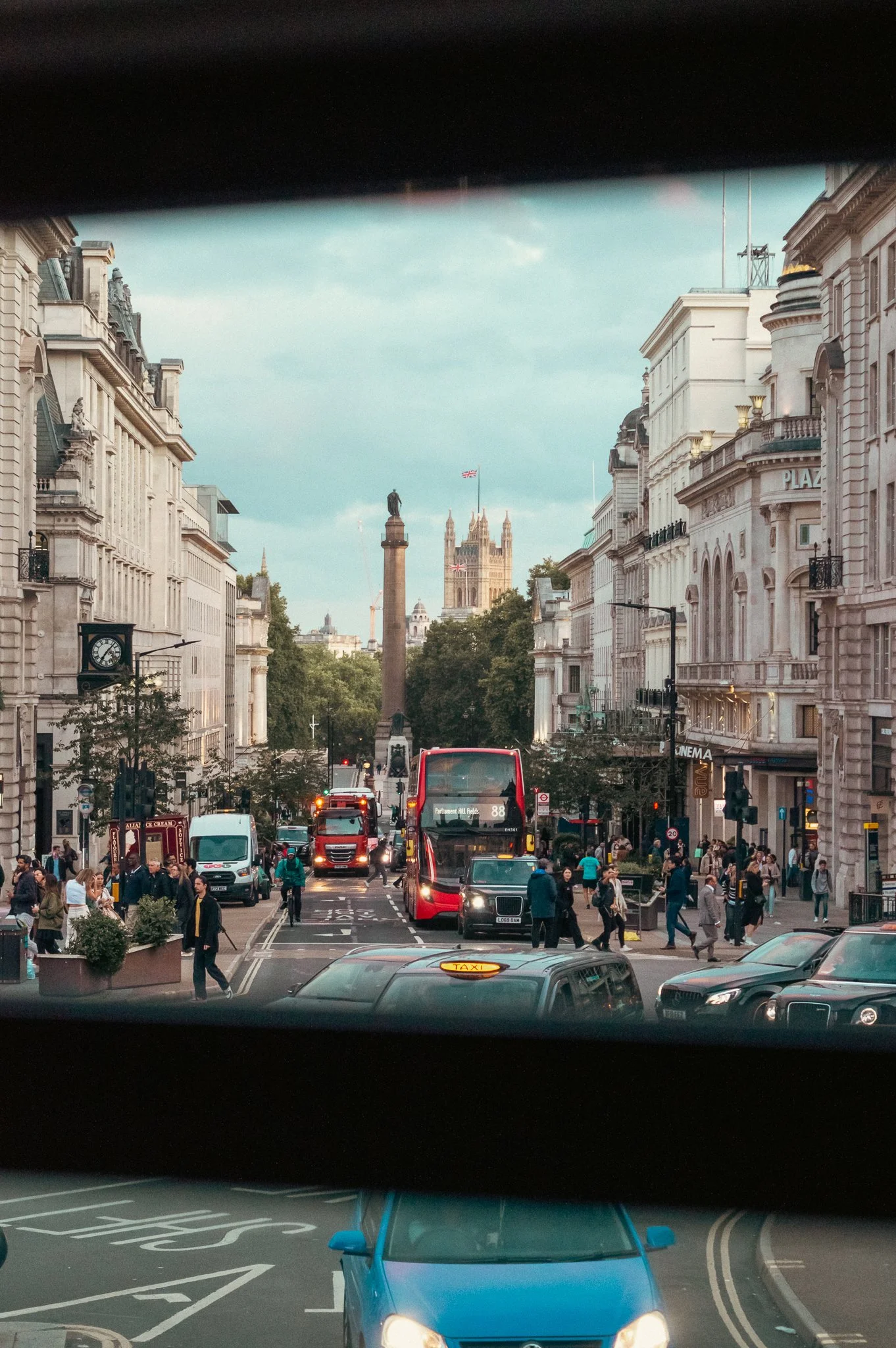 City street seen through a bus window, with buildings on both sides, a clear view of Big Ben in the distance, and a busy intersection with cars, buses, and pedestrians.