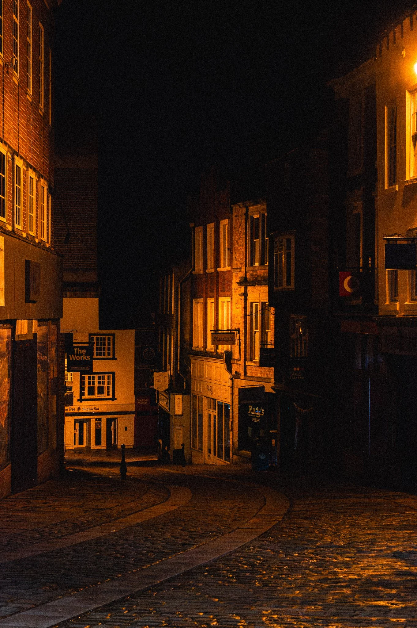 Nighttime street scene in a small town with cobblestone pavement, illuminated buildings, and signs on storefronts.