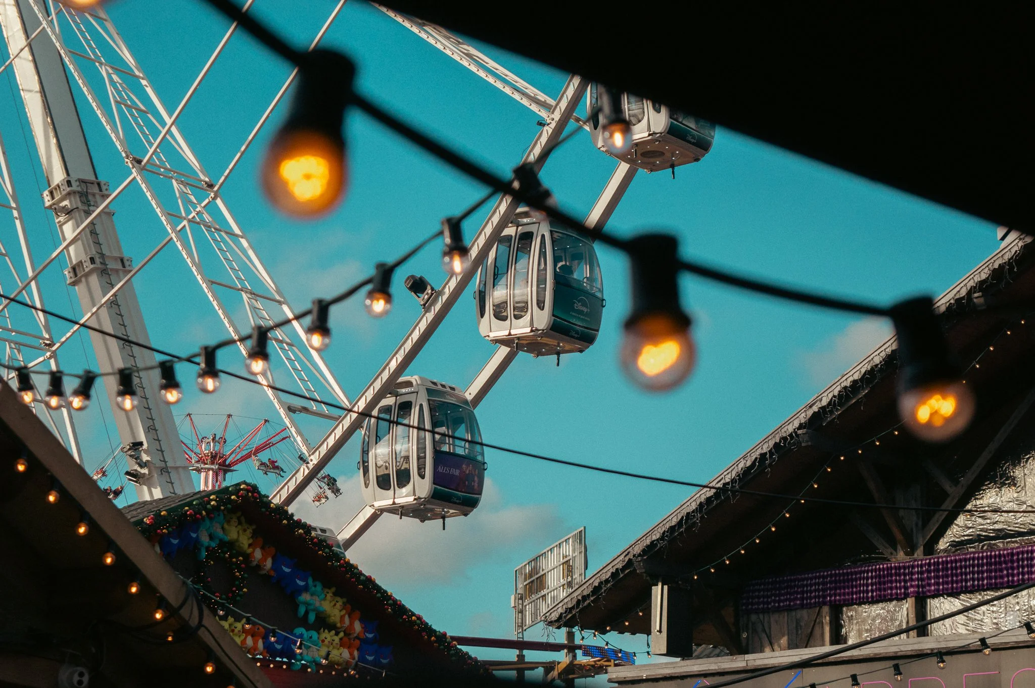 A Ferris wheel at an outdoor fair with a clear blue sky, festive lights, and colorful decorations beneath it.