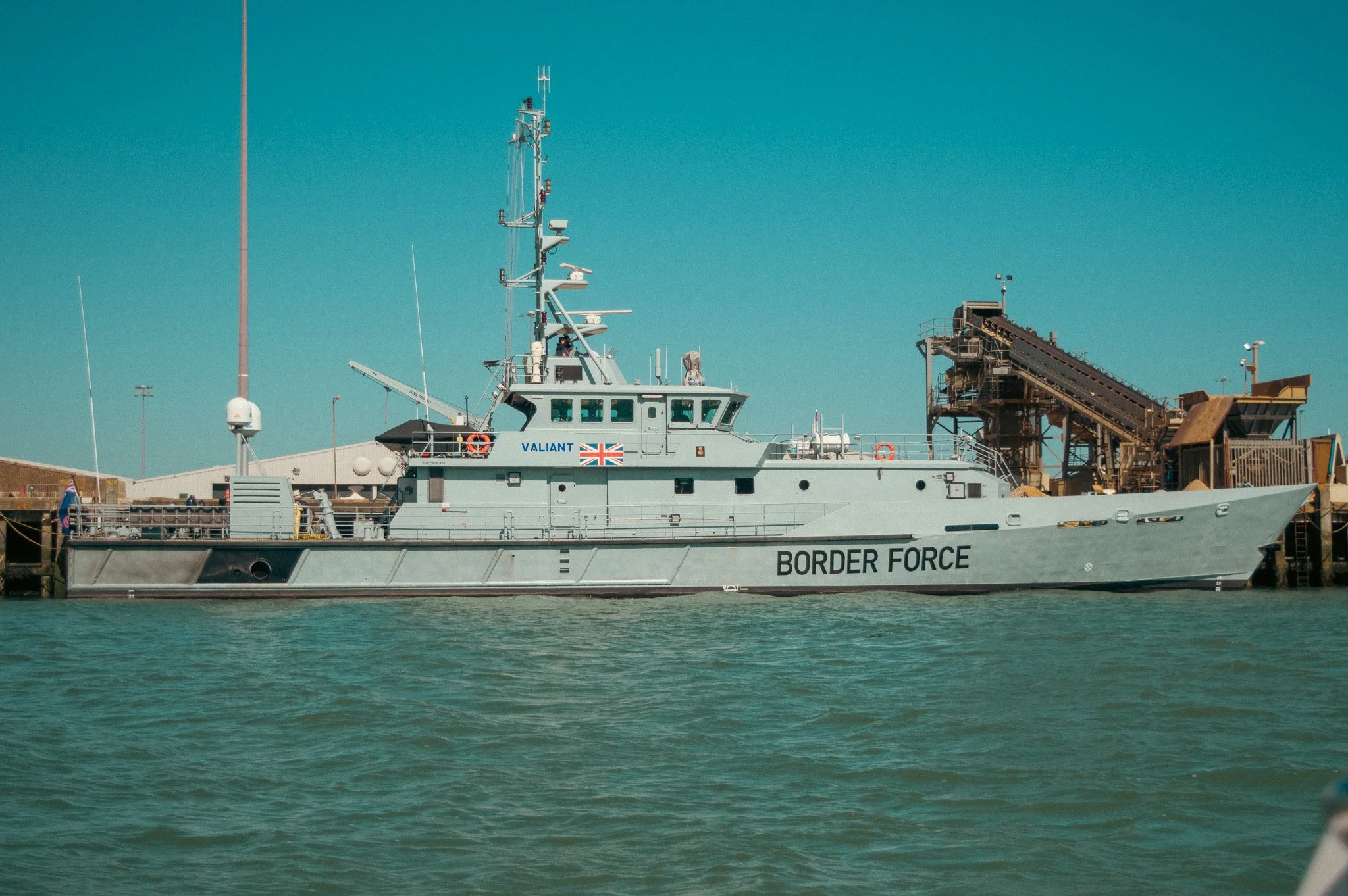 A Border Force patrol boat named Valiant docked at a harbor, with a clear blue sky in the background and industrial equipment nearby.
