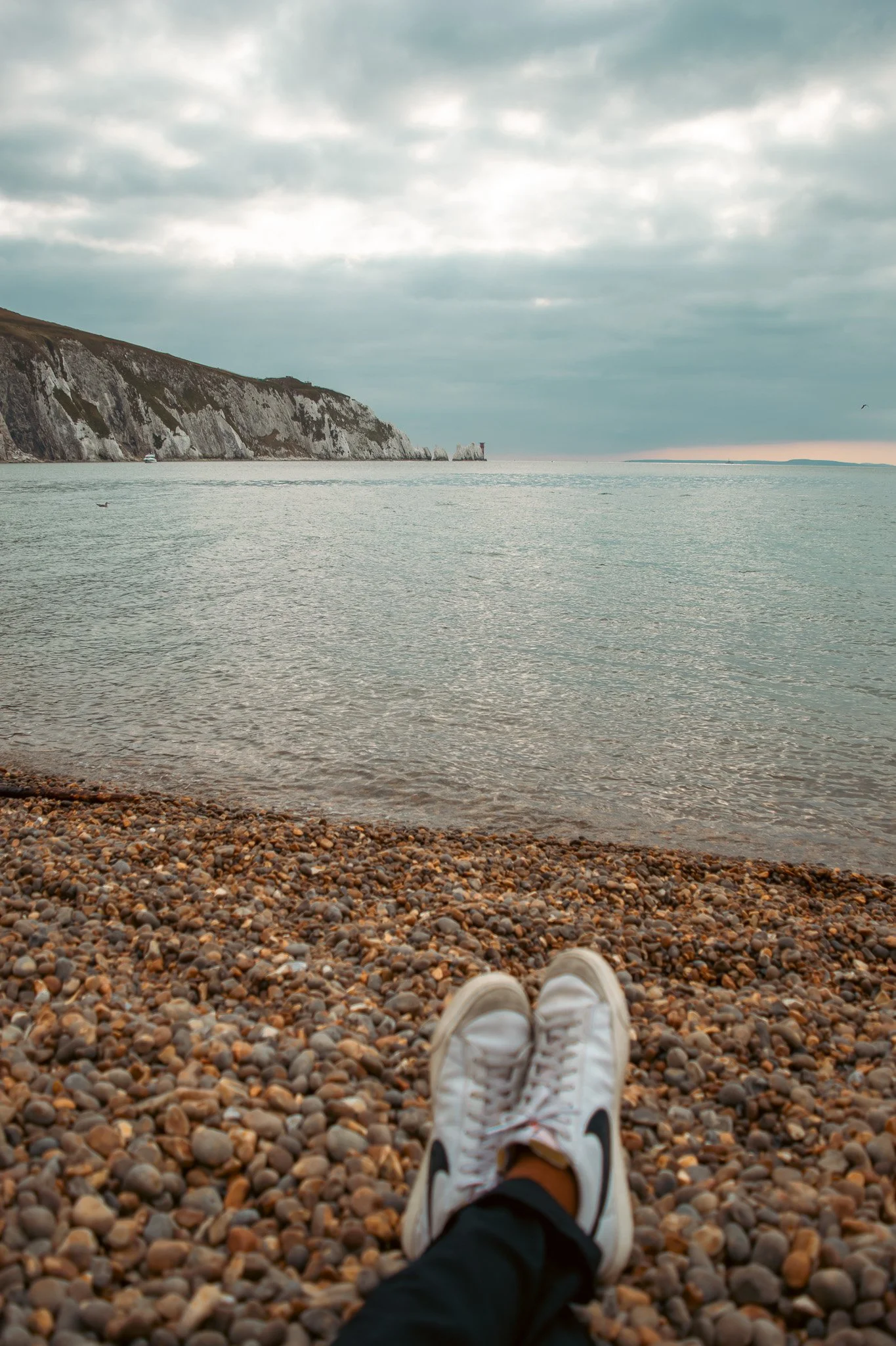 View from someone's perspective sitting on a pebble beach with legs extended, wearing white sneakers, looking out over the water towards distant cliffs and a lighthouse on a cloudy day.