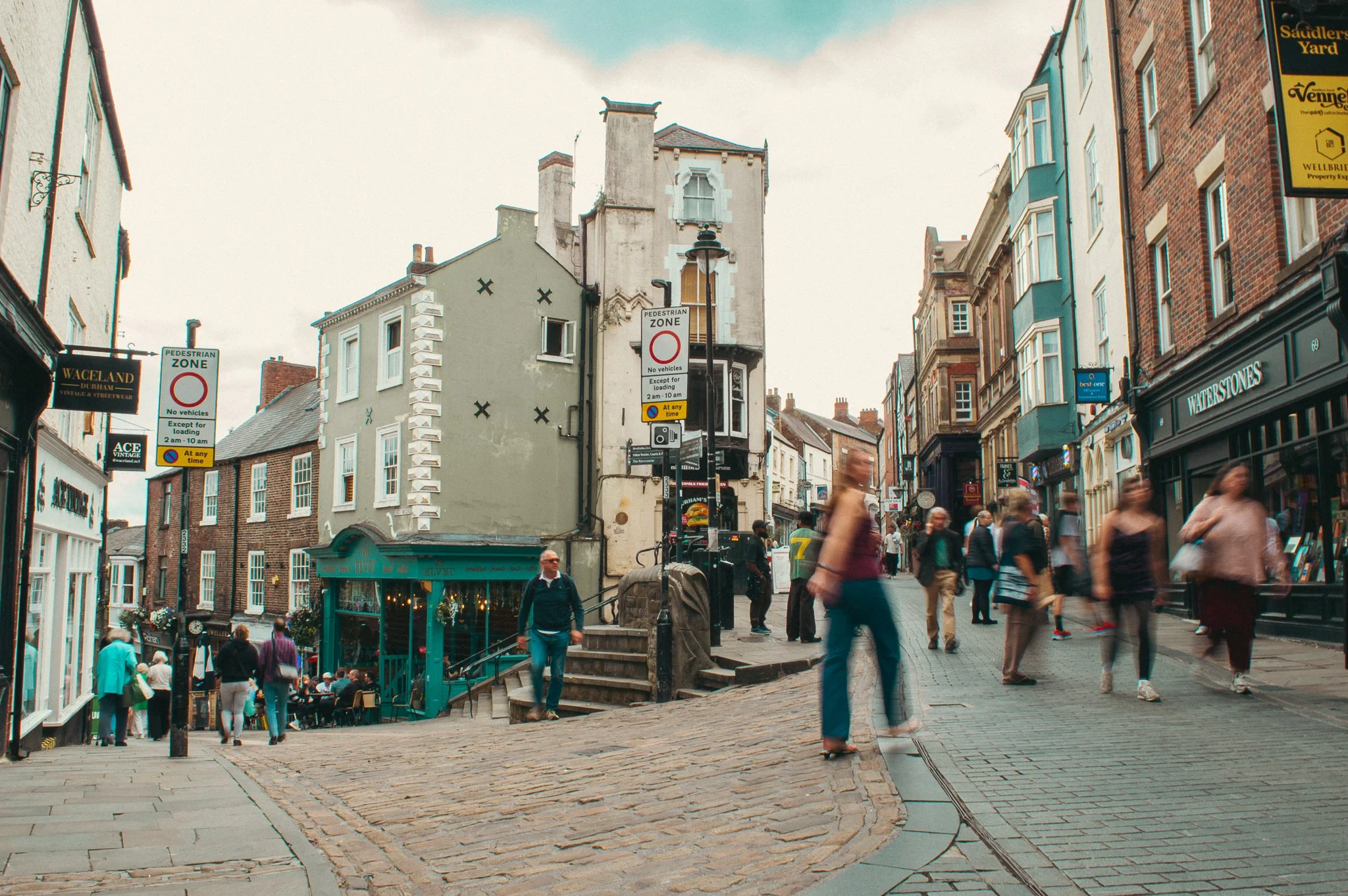 A bustling pedestrian street in a historic city with shops, cafes, and old buildings, with people walking and sitting outside, and a store with a sign that reads 'WATSTONES'.