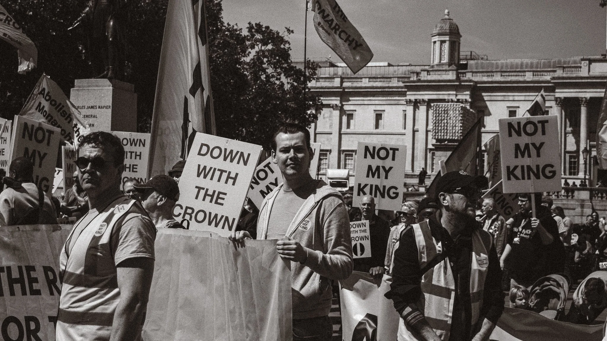 Protesters holding signs that read "DOWN WITH THE CROWN" and "NOT MY KING" outside a government building, with trees and a domed structure in the background.