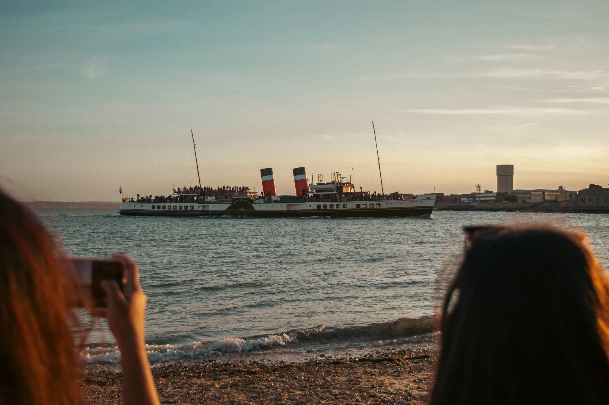 A large vintage steamship with two red and black smokestacks in the water, viewed from the beach with people taking photos, during sunset.