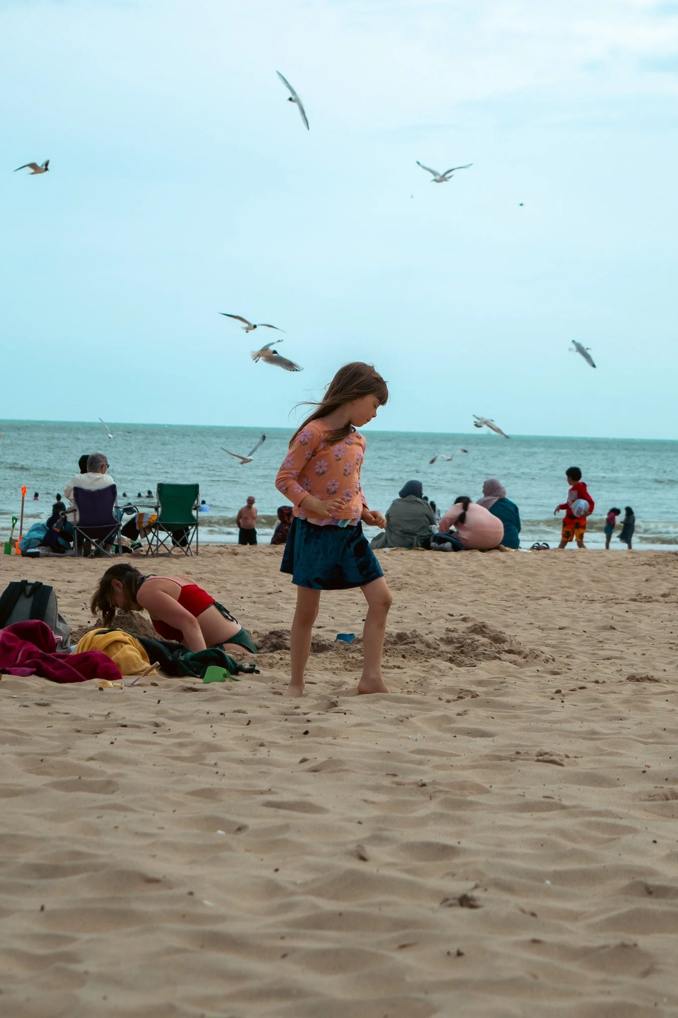 A girl walking on the sandy beach near the ocean, with seagulls flying overhead and people relaxing by the water.