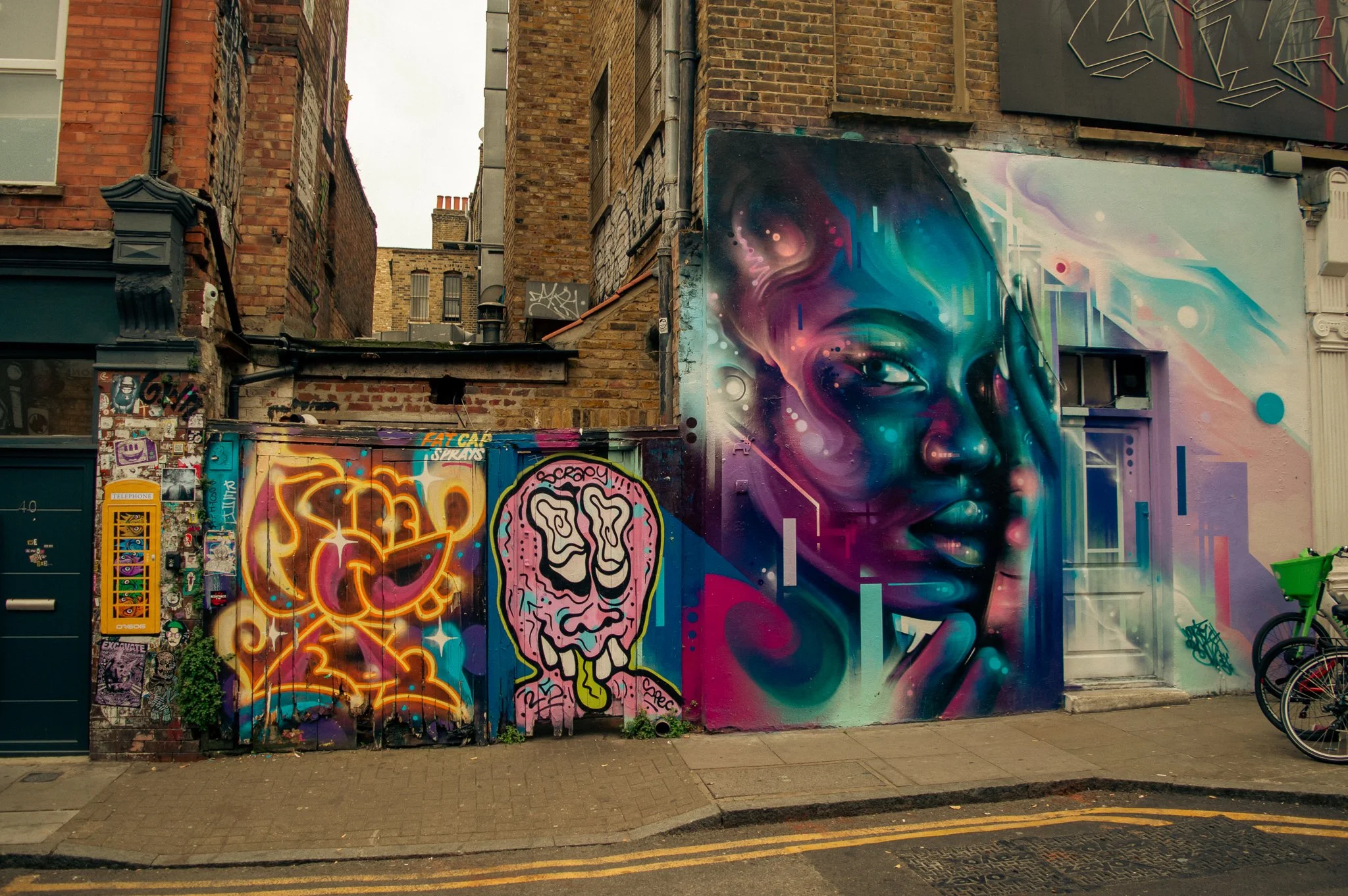 Street wall with colorful graffiti art, including a large realistic portrait of a woman with her eyes closed and hand on her face, a stylized face with brain and lightning motifs, and various tags and stickers, set against brick buildings in an urban