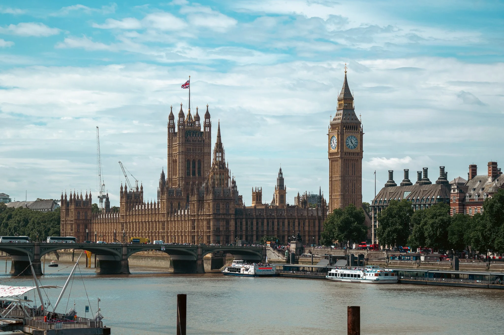 View of the Westminster Palace and Big Ben in London, with boats on the River Thames in the foreground and a partly cloudy sky overhead.