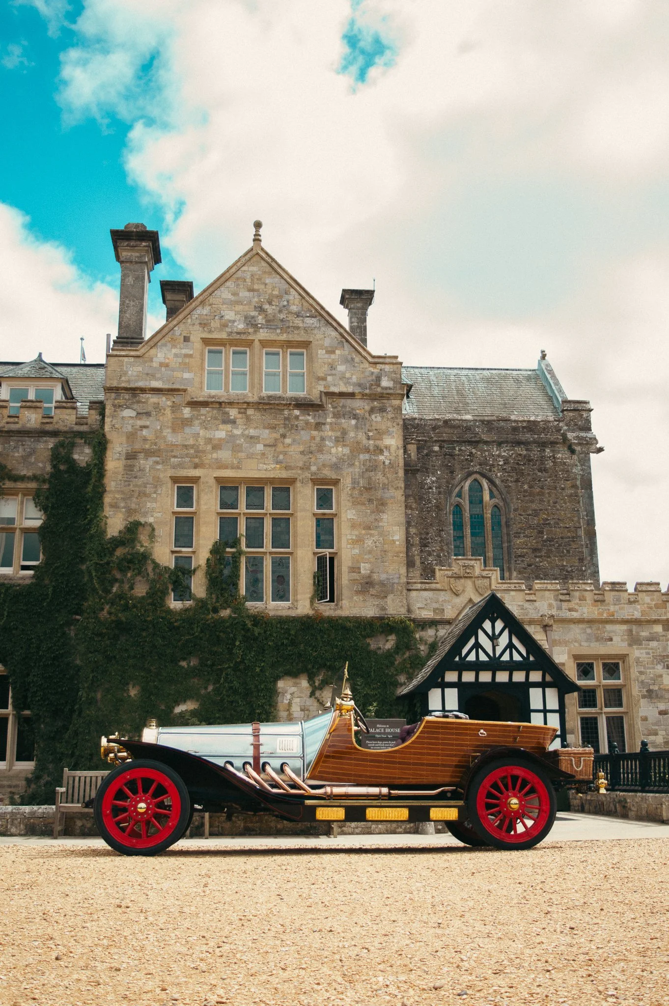 A vintage car with wooden paneling and red wheels parked in front of an old stone building with Gothic windows and ivy climbing the walls.