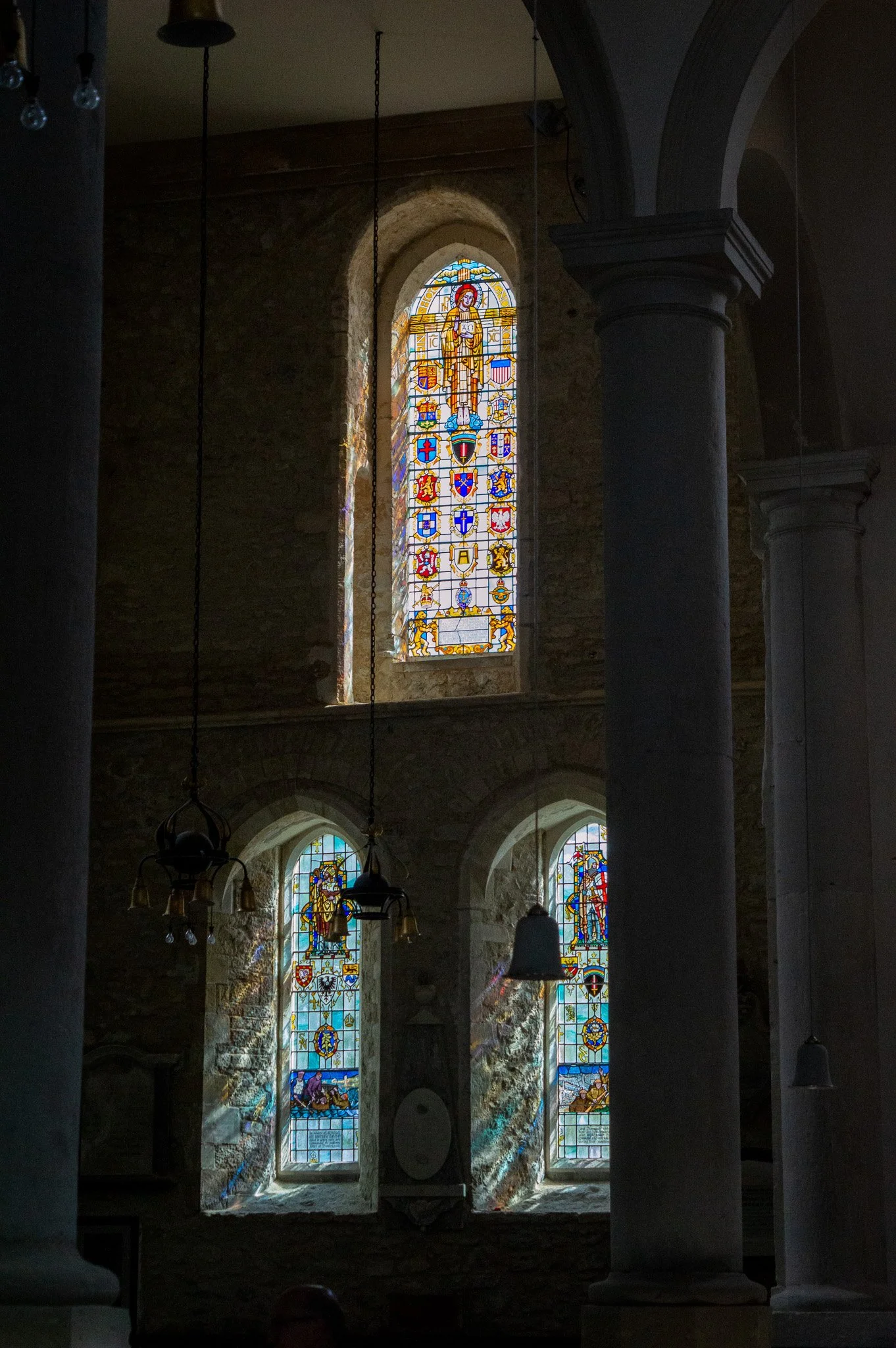 Inside a stone church with stained glass windows depicting religious scenes and saints, with pillars and hanging chandeliers.