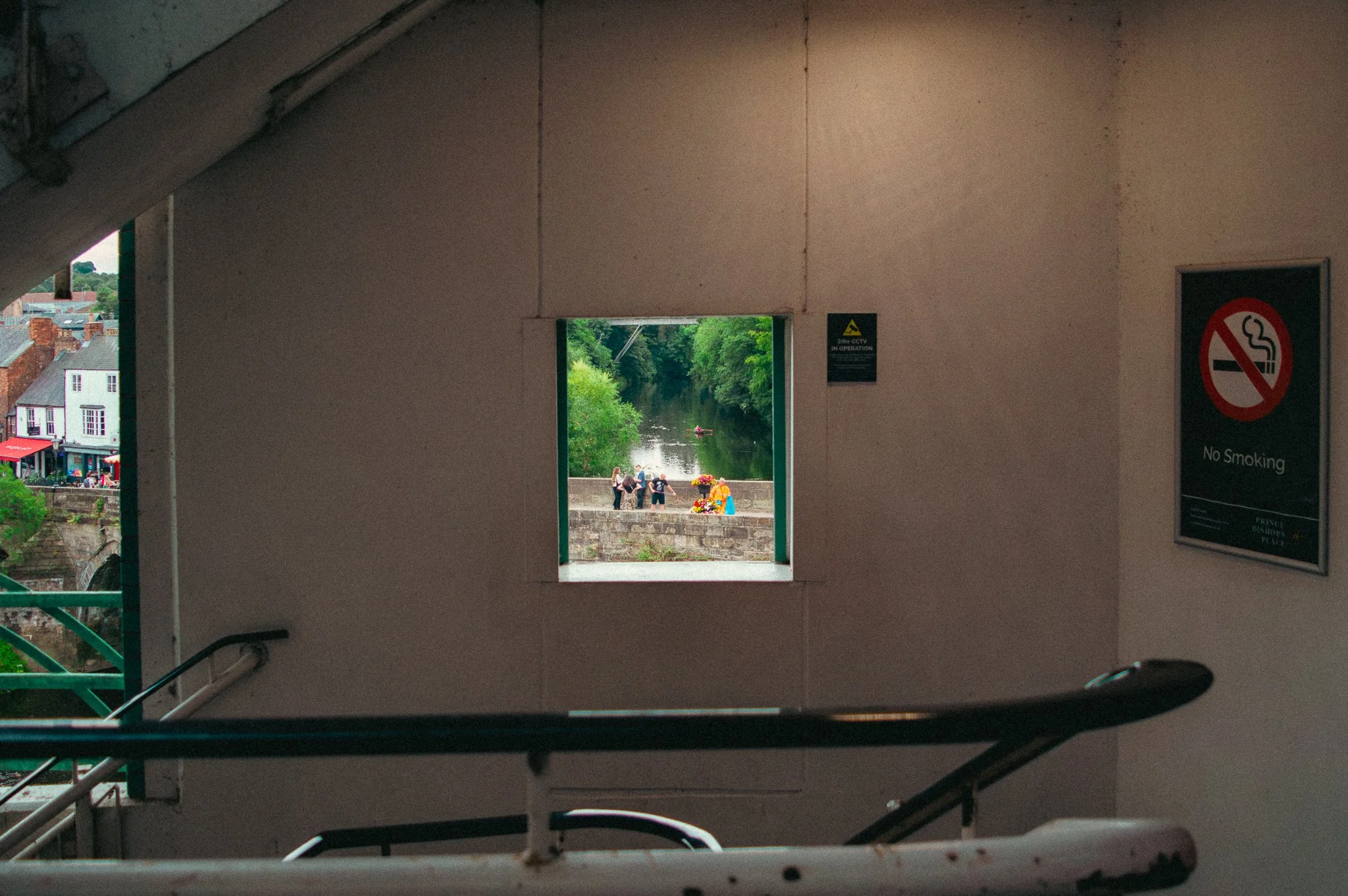 View through a small window in a building showing people gathered near a river with trees and buildings in the background. The photo is taken from inside a parking garage or similar structure.