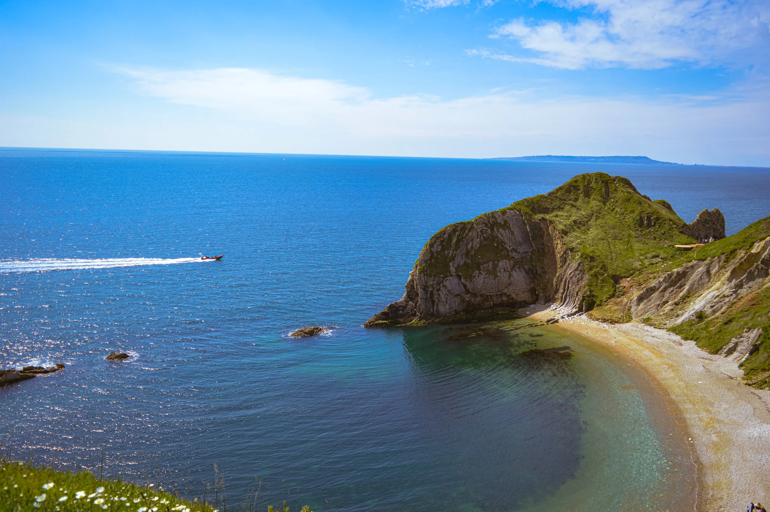 A coastal landscape featuring a blue ocean with a boat passing by, green cliffs, a small beach, and a partly cloudy sky.