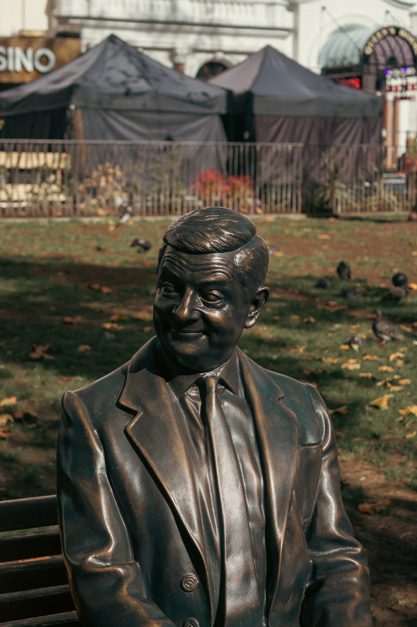 Bronze bust of a man wearing a suit and tie, set outdoors with tents and a building in the background, with falling leaves on the ground.