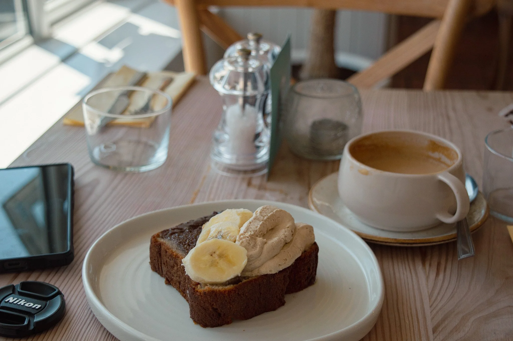 A white plate with a slice of banana bread topped with banana slices. In the background, a wooden table with various glasses, a coffee cup, and a salt and pepper shaker.