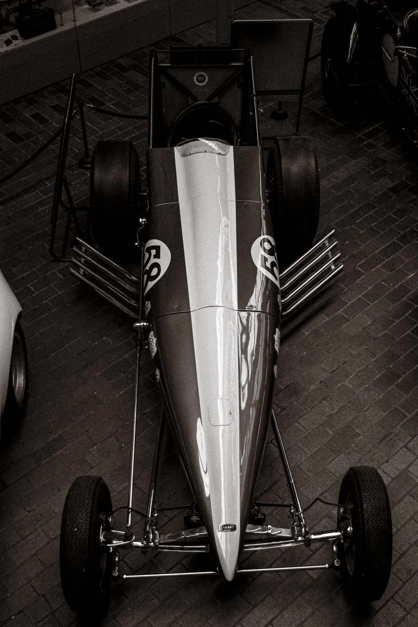A vintage race car with a sleek, polished body and large tires, displayed indoors on a brick floor. The car has racing decals and a minimalistic cockpit design.