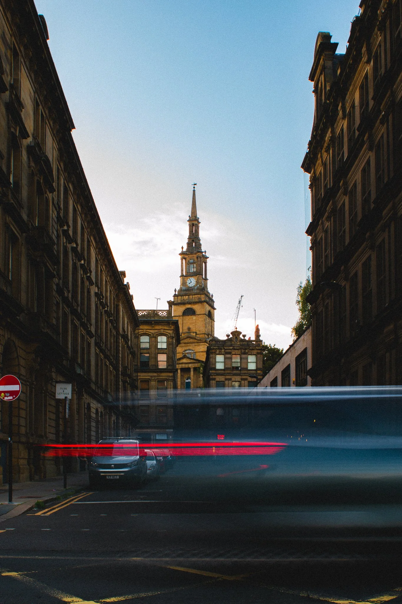 Street view with historic church tower, parked cars, and buildings on both sides under a clear sky at dusk.